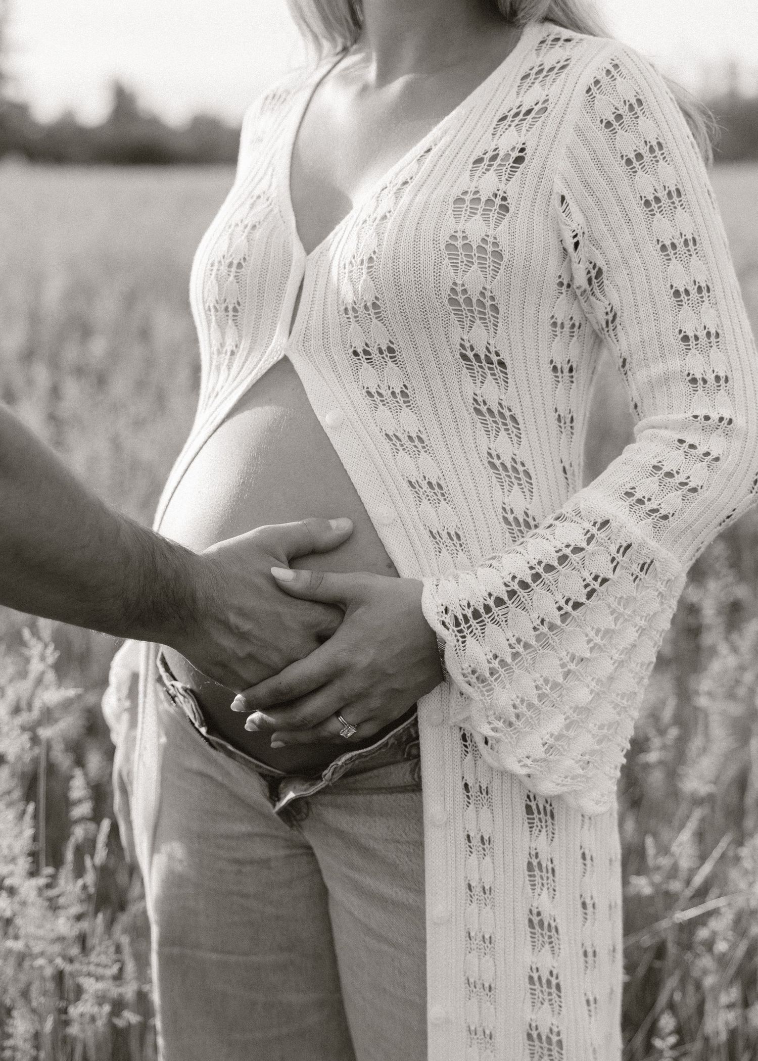 Black and white maternity photo of pregnant woman in knit cardigan with partner's hands cradling baby bump in field.