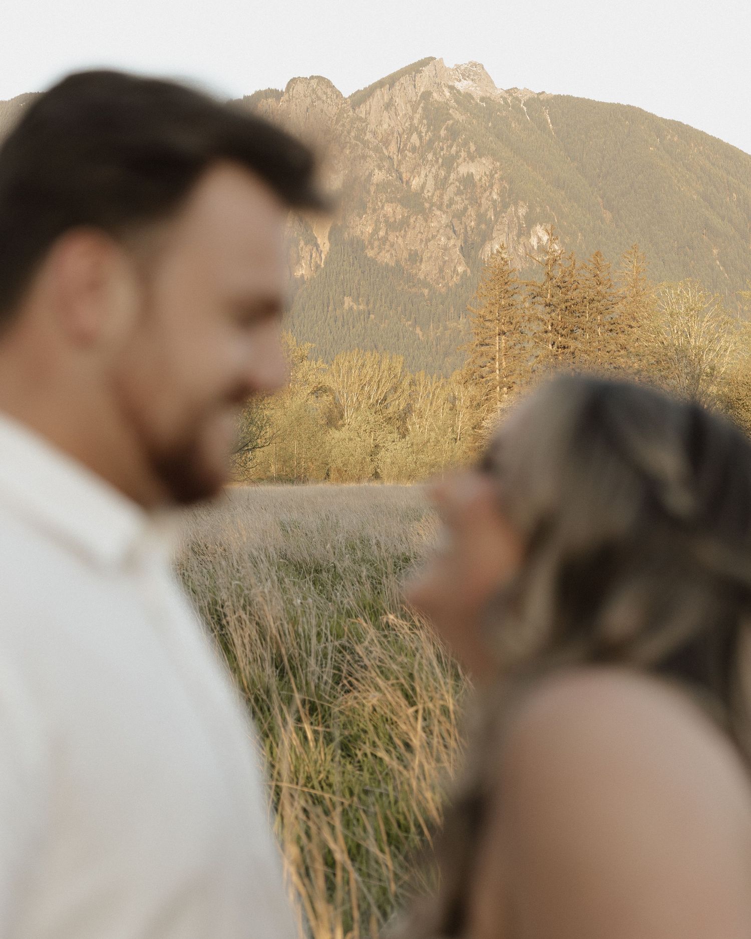 Romantic couple shares intimate moment in field with mountain landscape behind them.