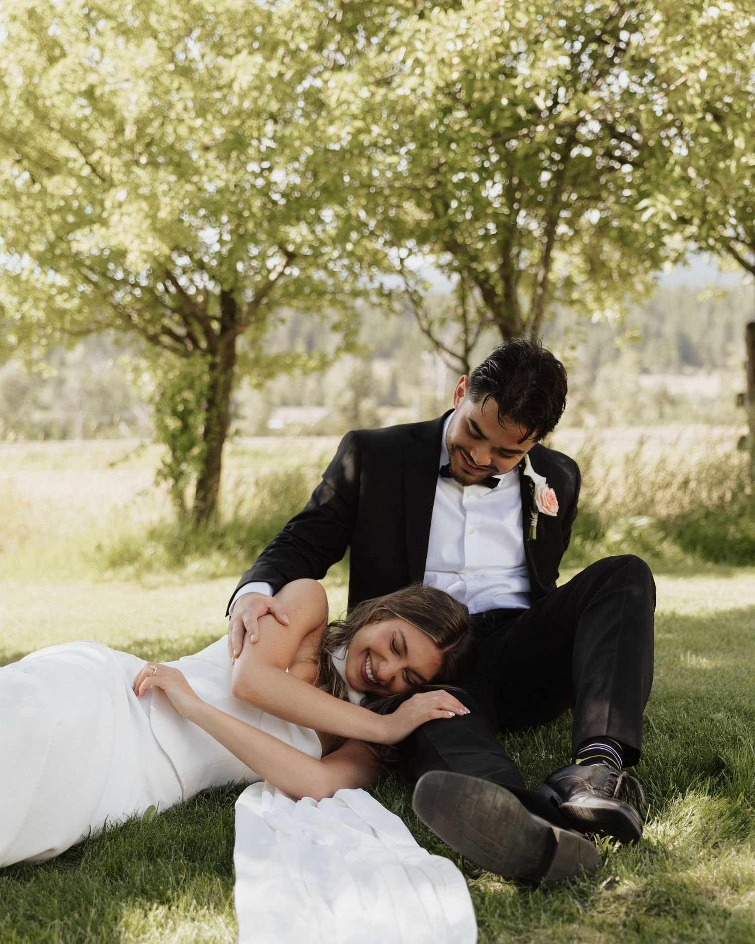 Newlyweds share a romantic moment lying in the grass under sunlit trees.