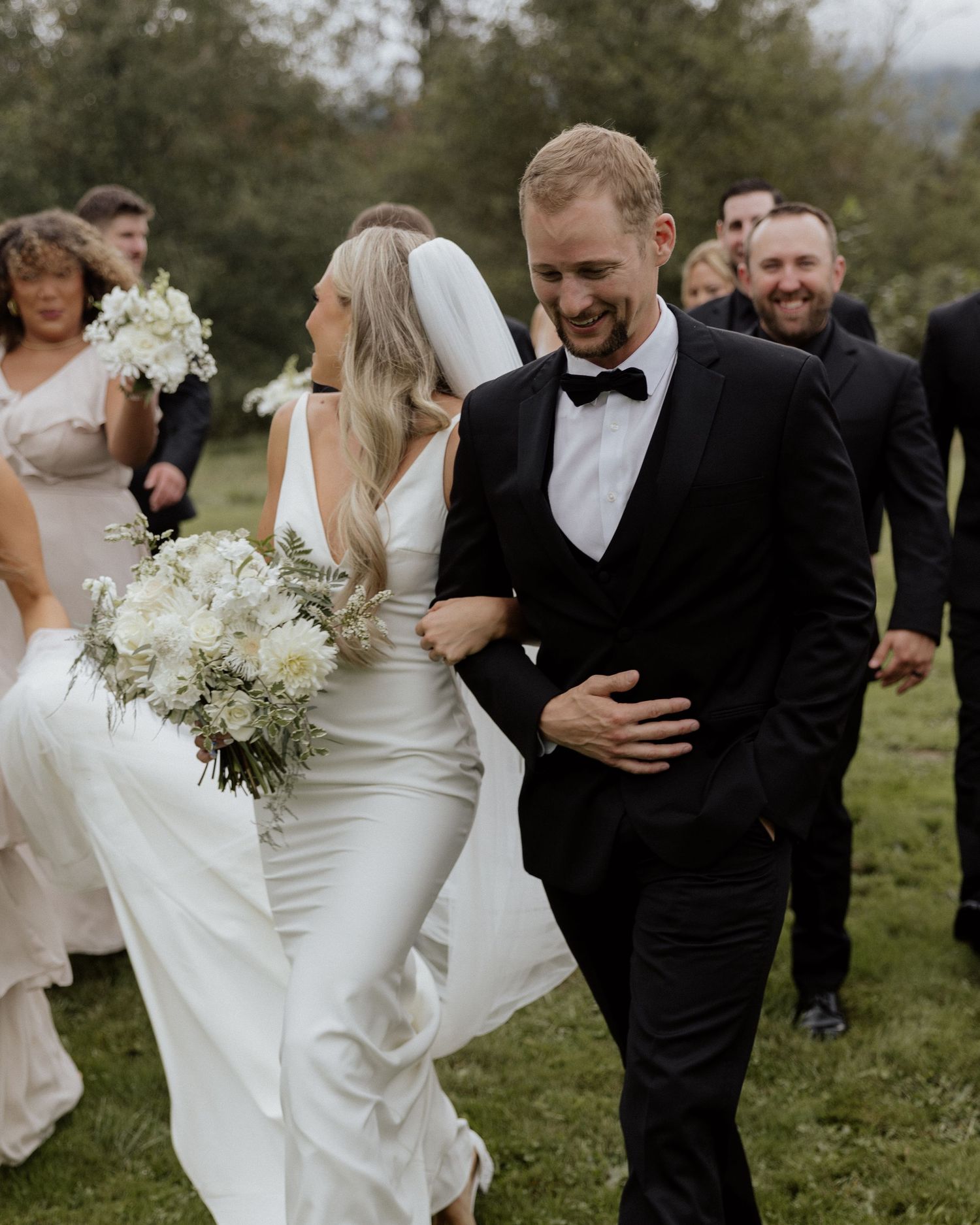 Joyful newlyweds walk with wedding party during outdoor celebration.