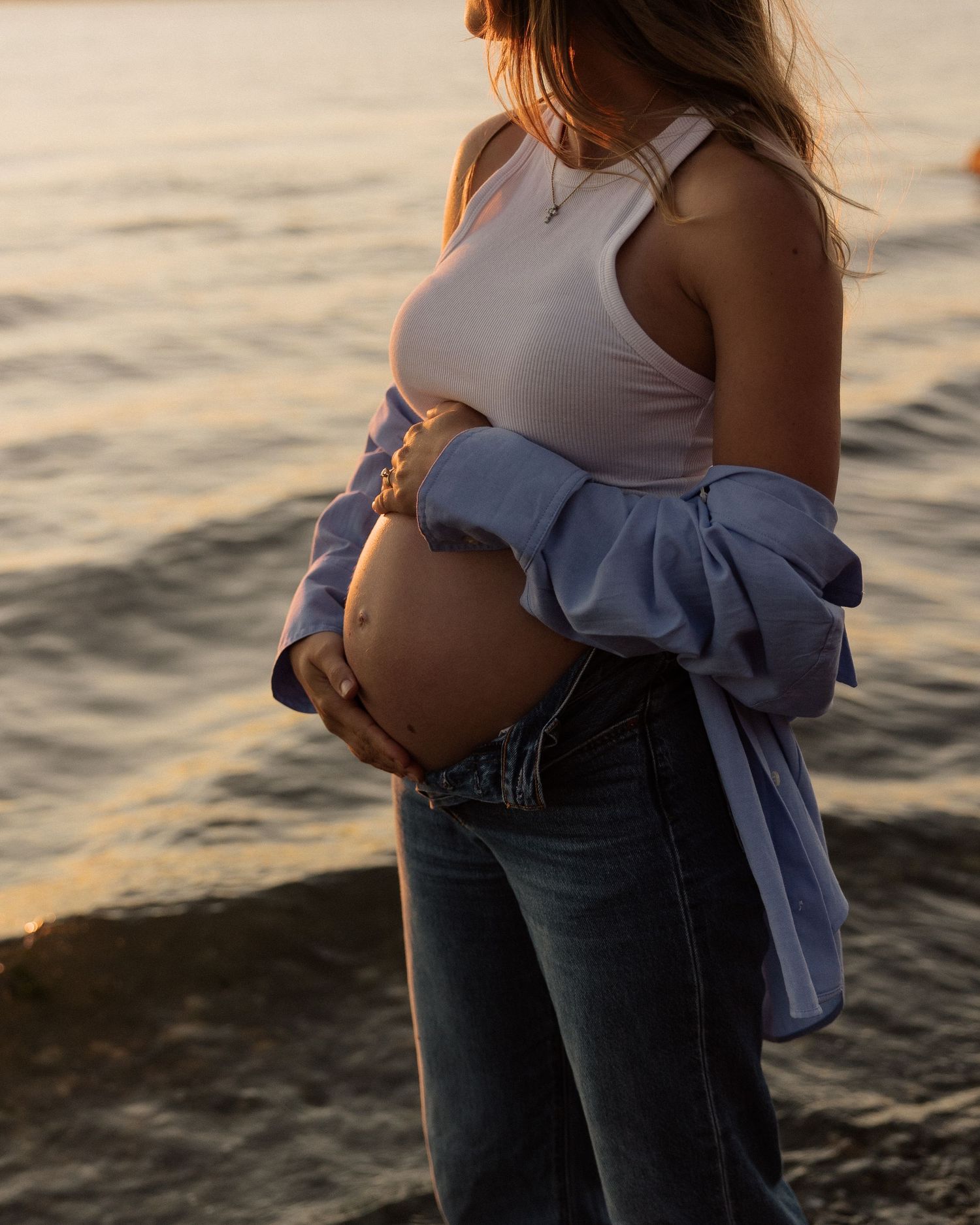 A close-up shot of an expectant mother's baby bump in white tank top and denim at the beach.