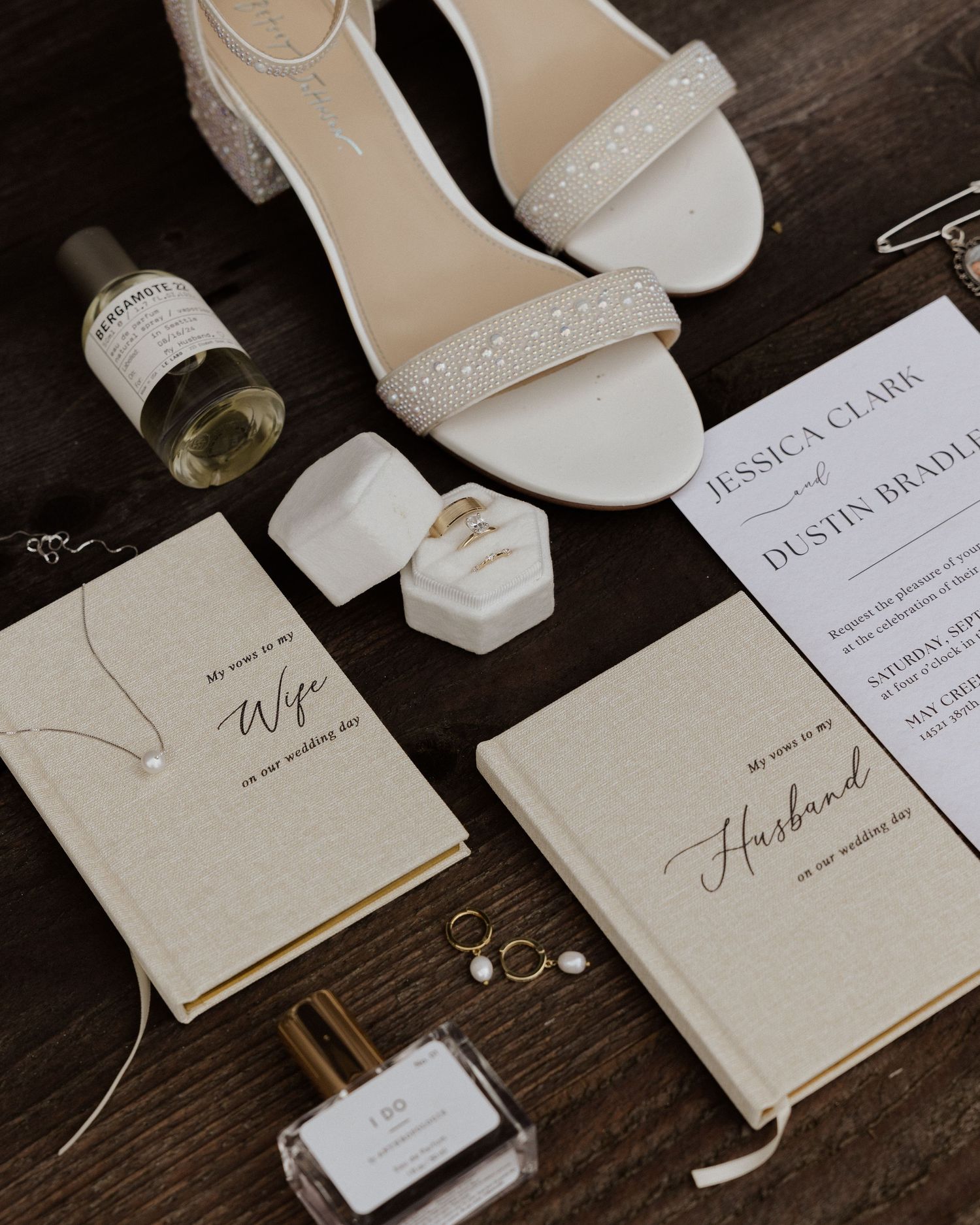 Elegant wedding accessories including white sandals, invitations and perfume arranged on wooden surface.