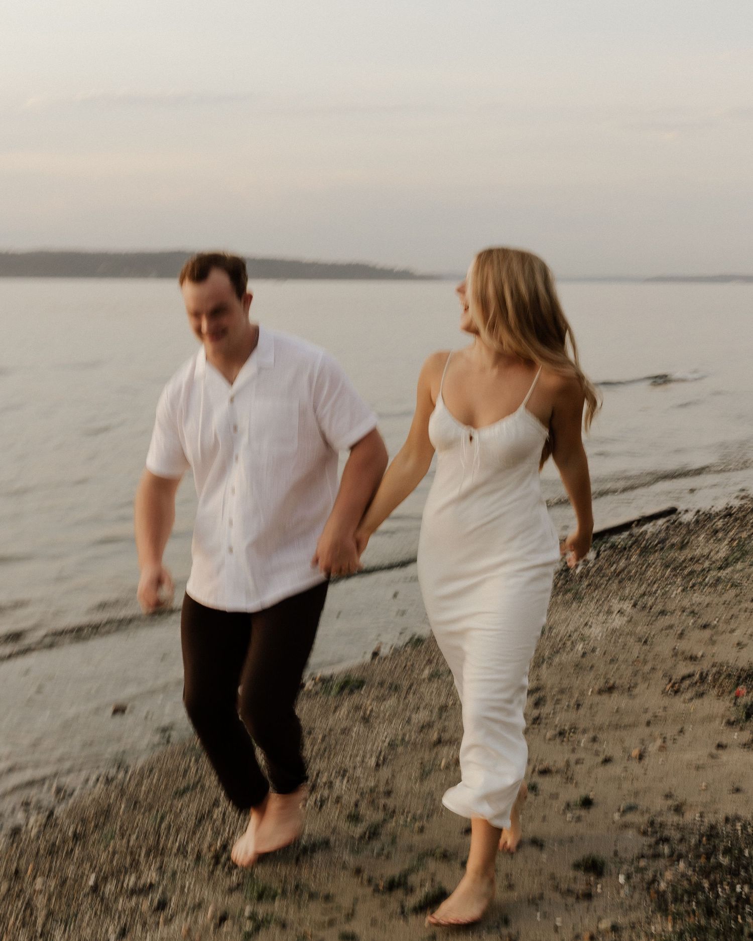 Two people in white outfits walk barefoot along the shoreline at dusk.