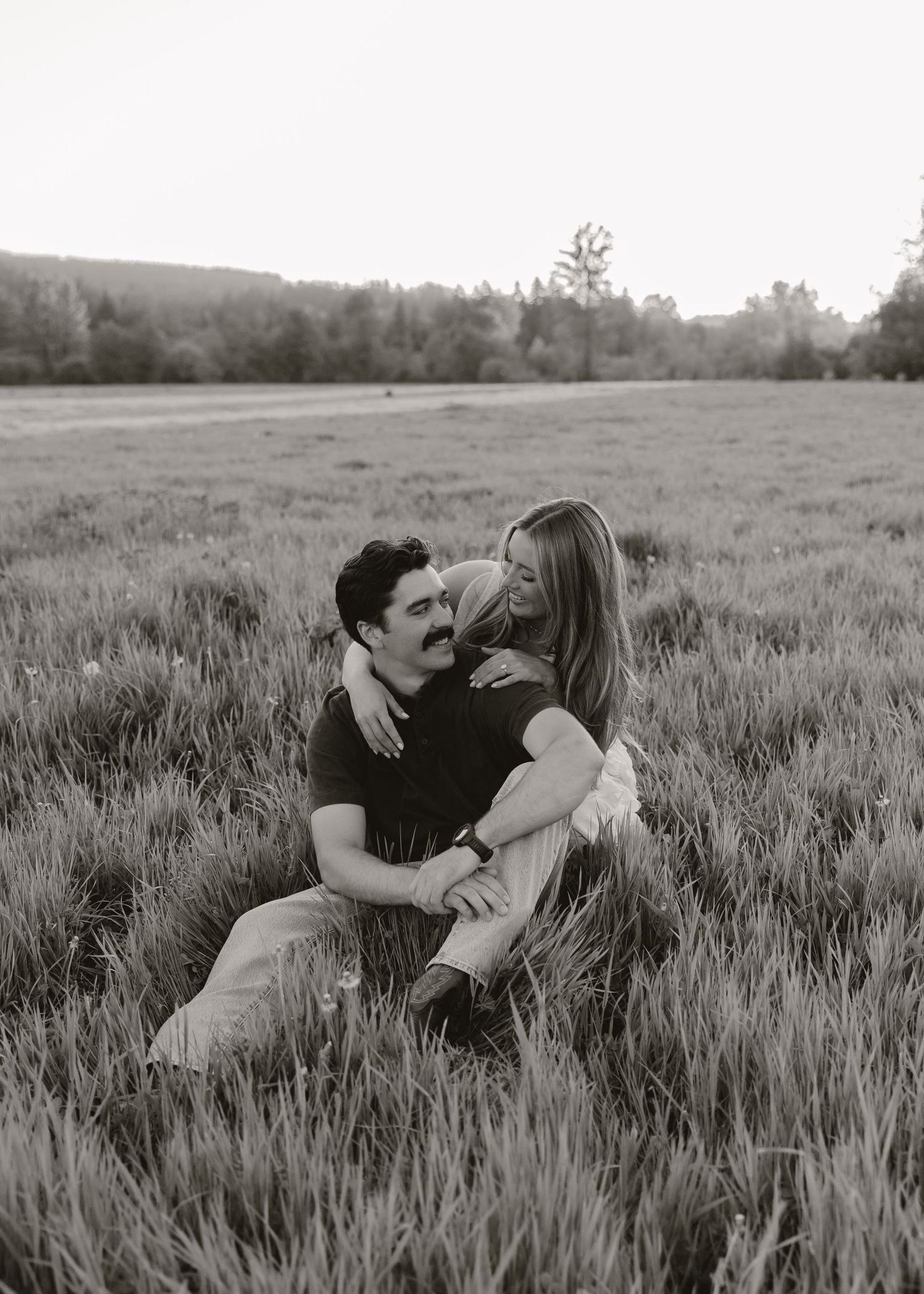 Black and white photo of a couple sitting in a tall grass field, sharing a tender embrace.