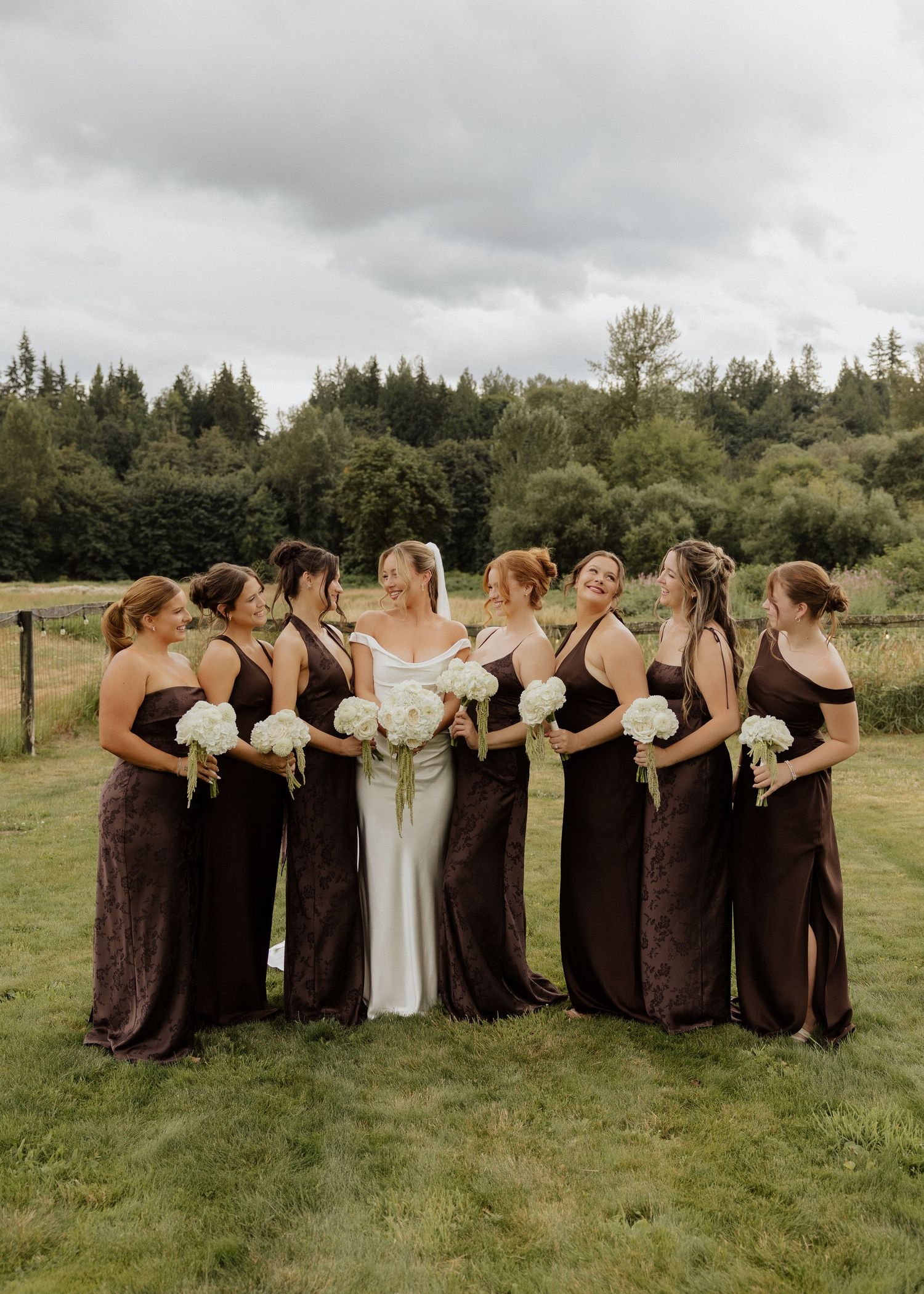 Bride in white gown with seven bridesmaids in dark brown dresses holding white bouquets outdoors.