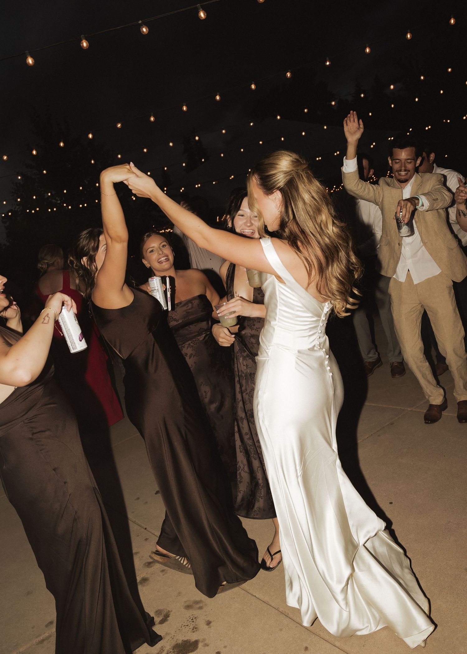 Bride in white satin gown dancing with guests under string lights at outdoor wedding reception.