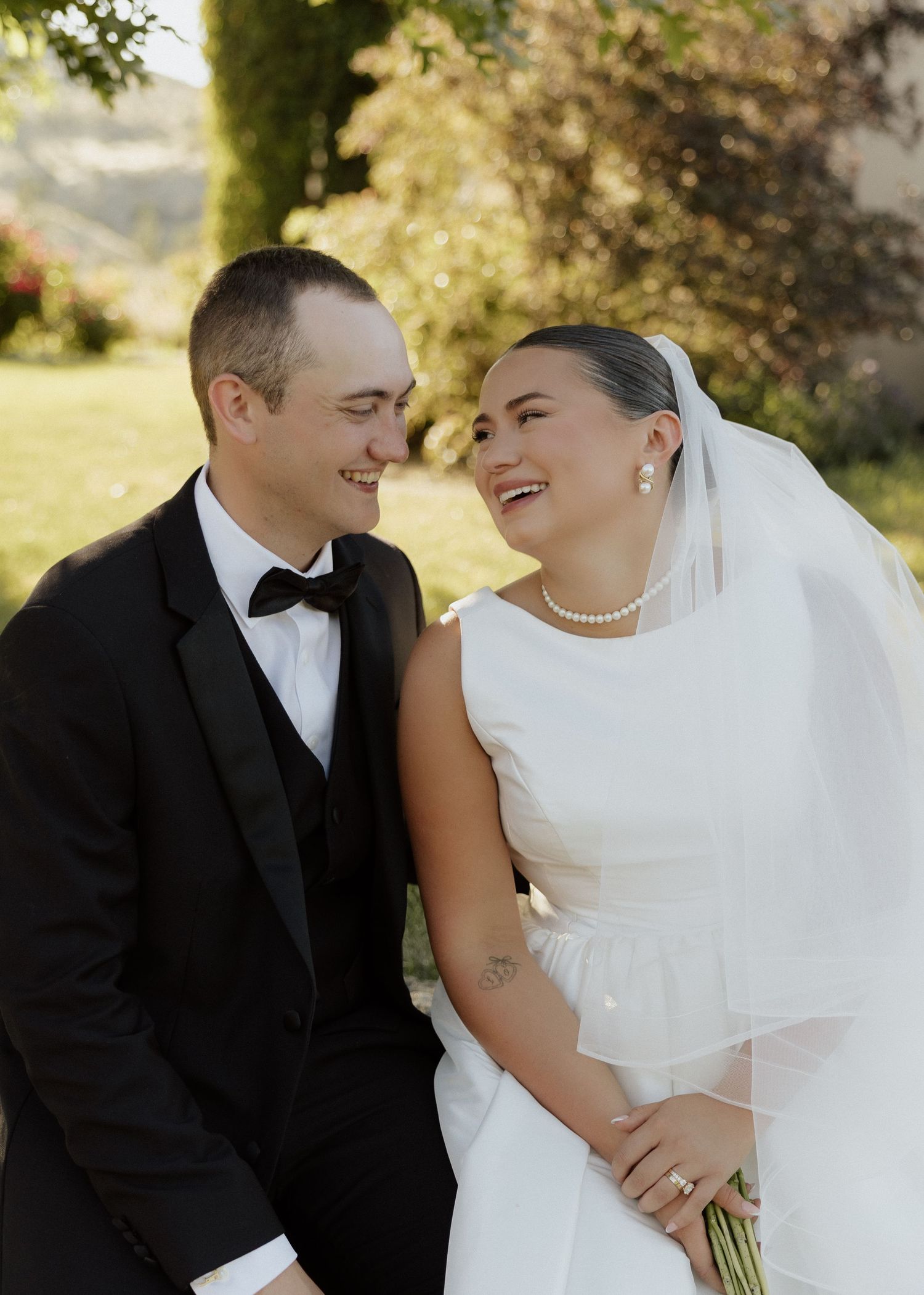 Smiling bride in white gown and veil with pearl necklace and groom in black tuxedo outdoors.