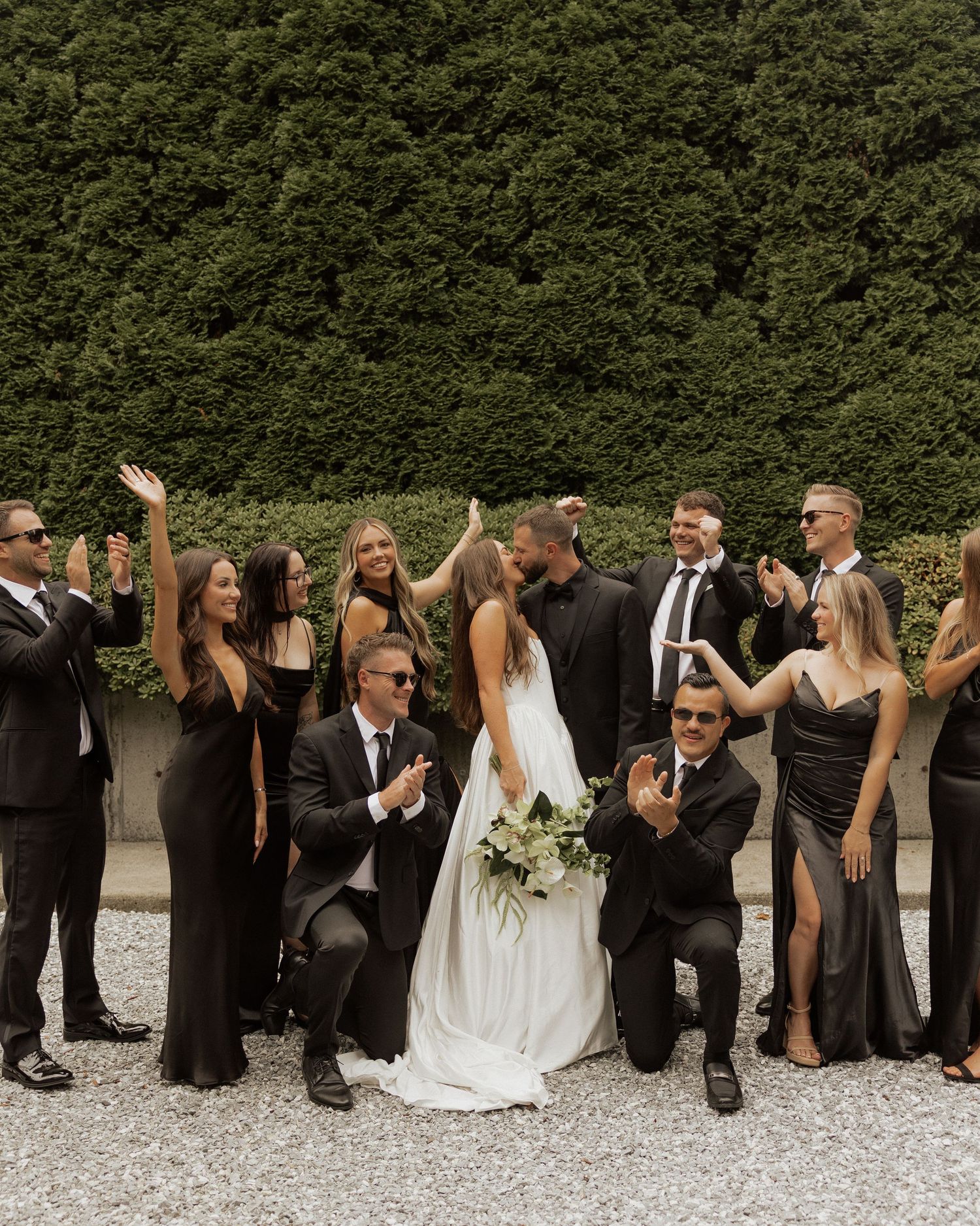 Wedding party in black attire celebrating kissing couple outdoors near hedge.