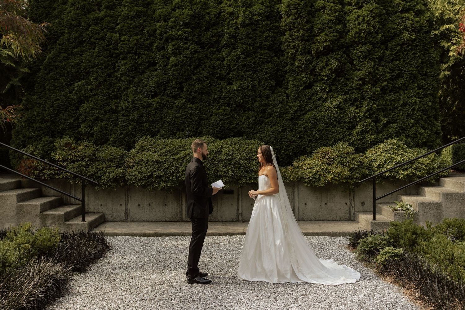 Bride and groom share a first look moment on a gravel path surrounded by tall green hedges.