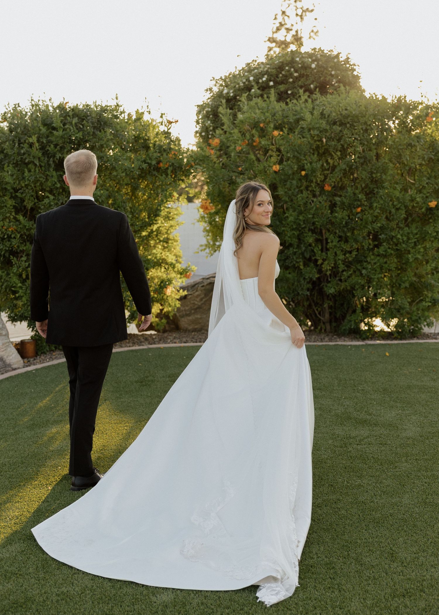 Bride in elegant white strapless gown with long train walks with groom in black tuxedo in garden.