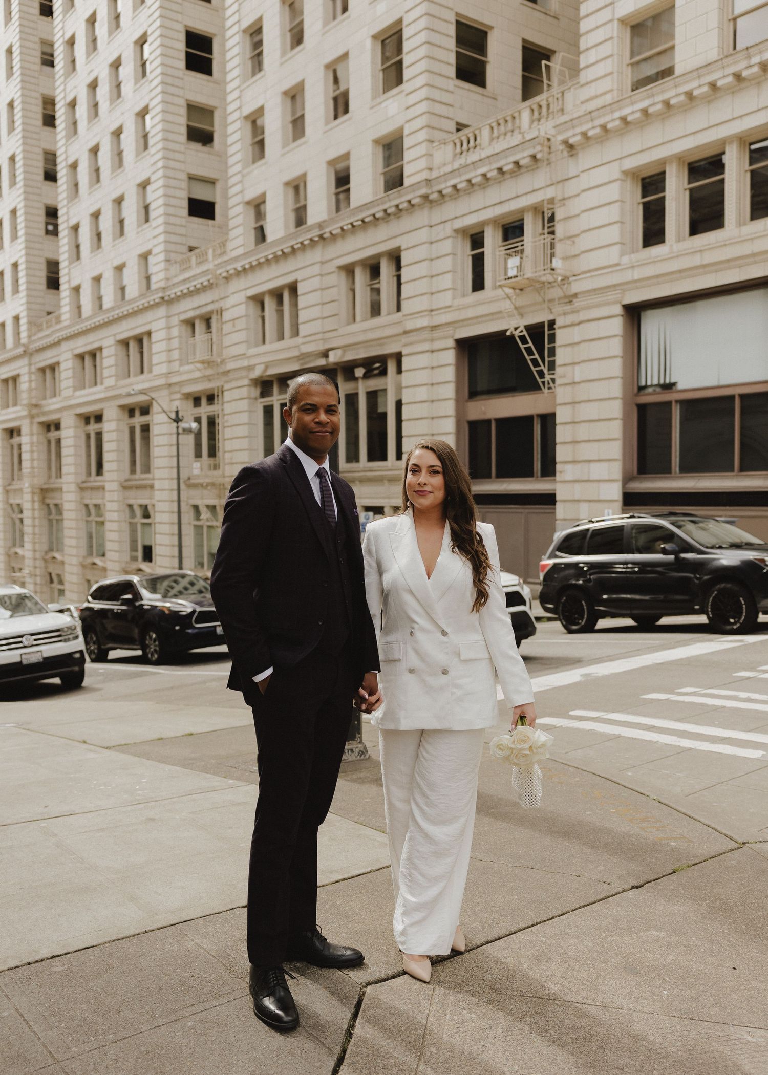 Elegant couple in matching white suit and dark tuxedo posing on a city sidewalk with classic architecture.