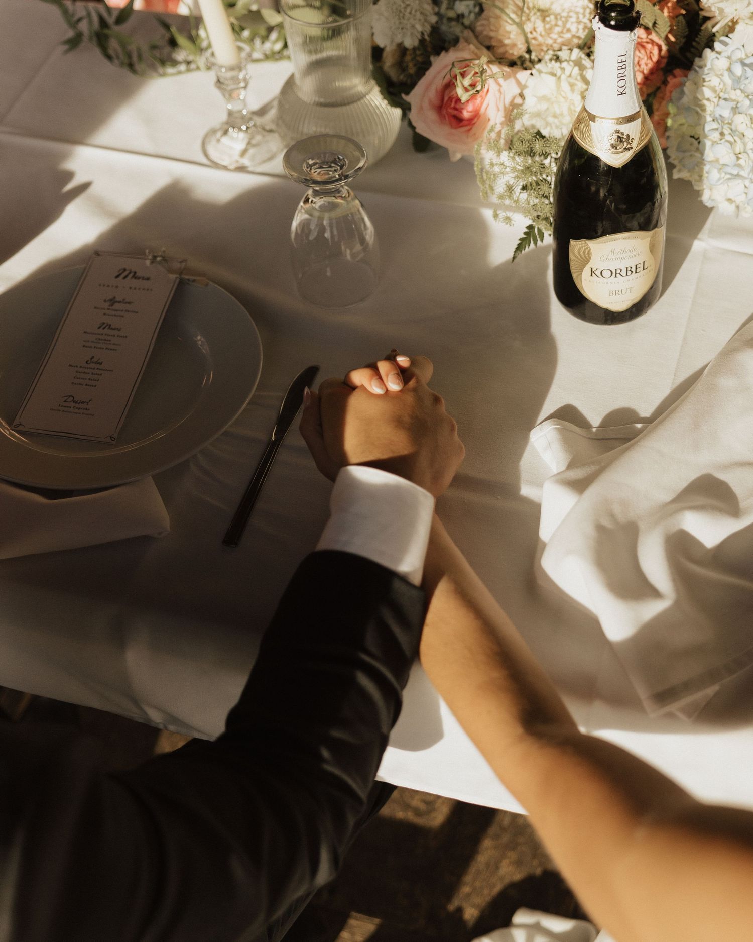 Couple holds hands at wedding reception table with champagne bottle and floral centerpiece.
