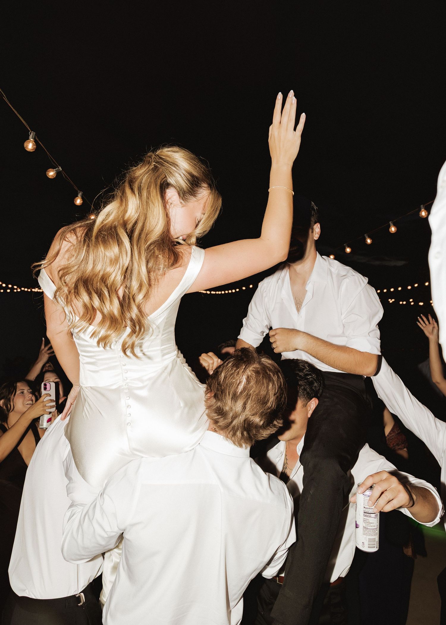 Bride in white satin gown lifted by wedding guests while dancing under string lights at nighttime reception.
