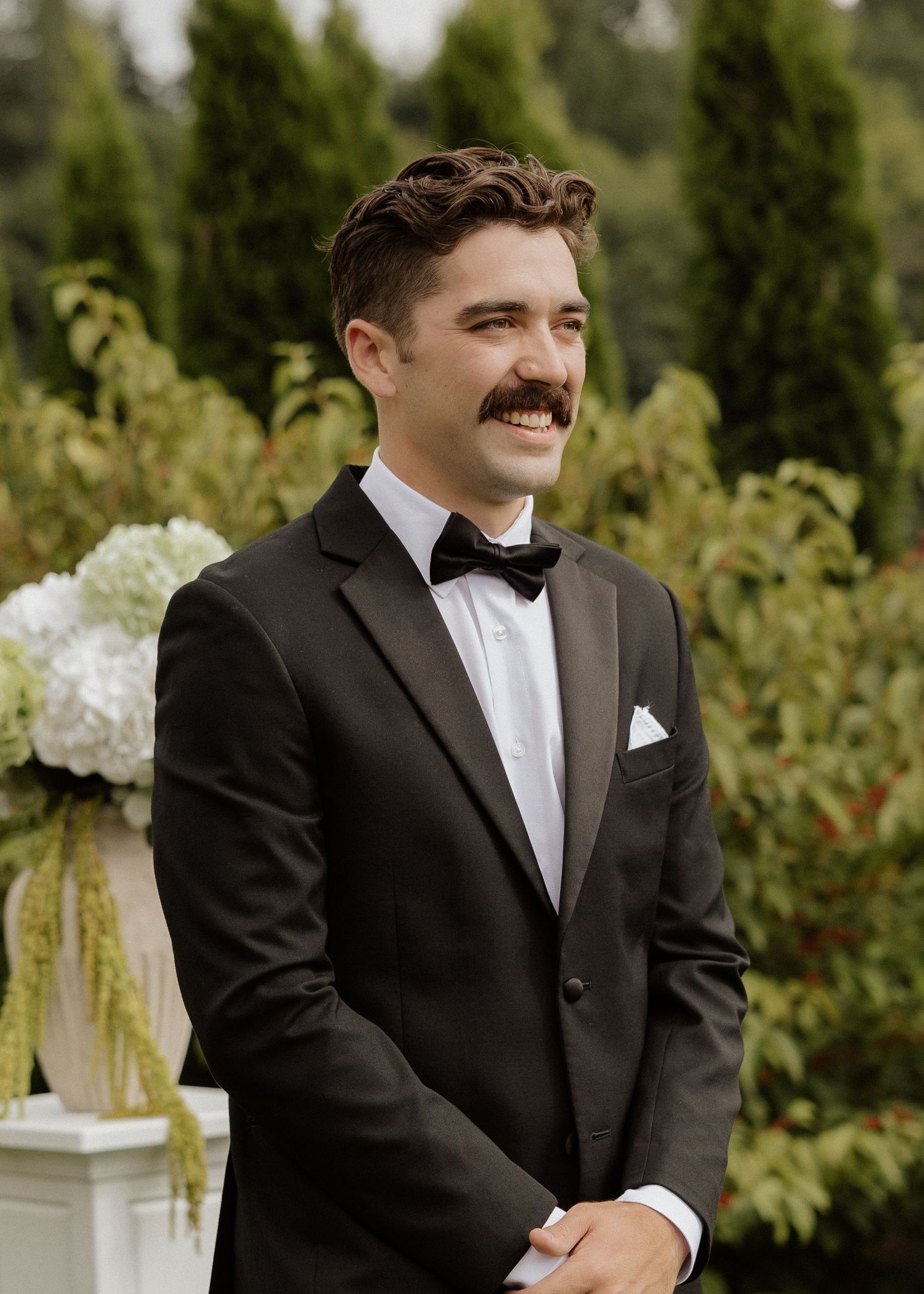 Groom in black tuxedo with bow tie and mustache smiling at outdoor wedding ceremony.