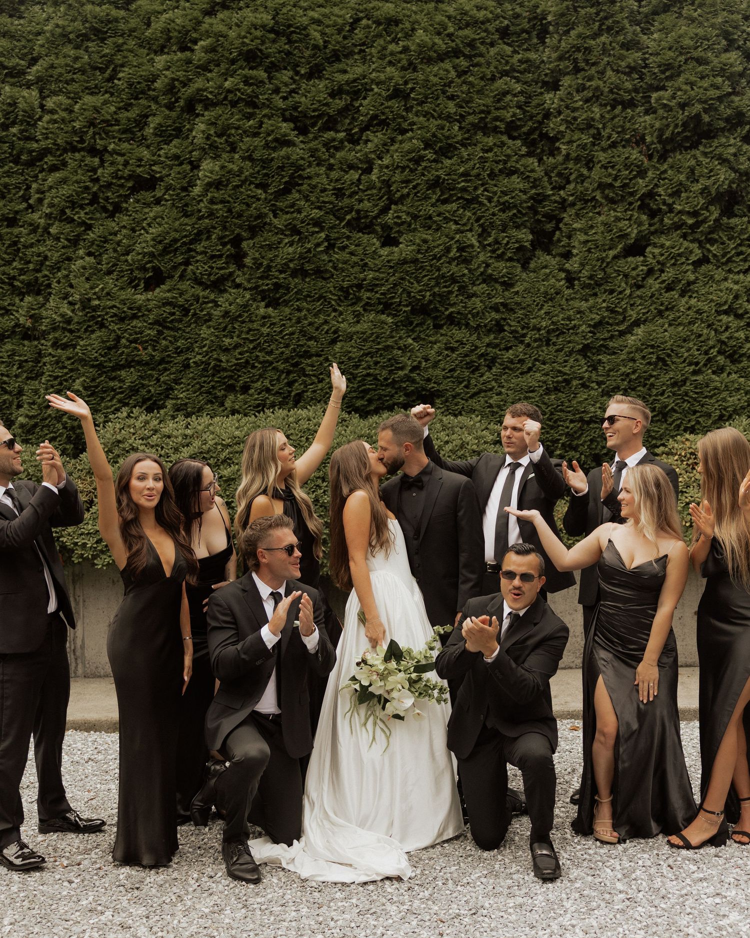 Wedding party in black attire celebrates with bride and groom kissing, surrounded by cheering guests outdoors.