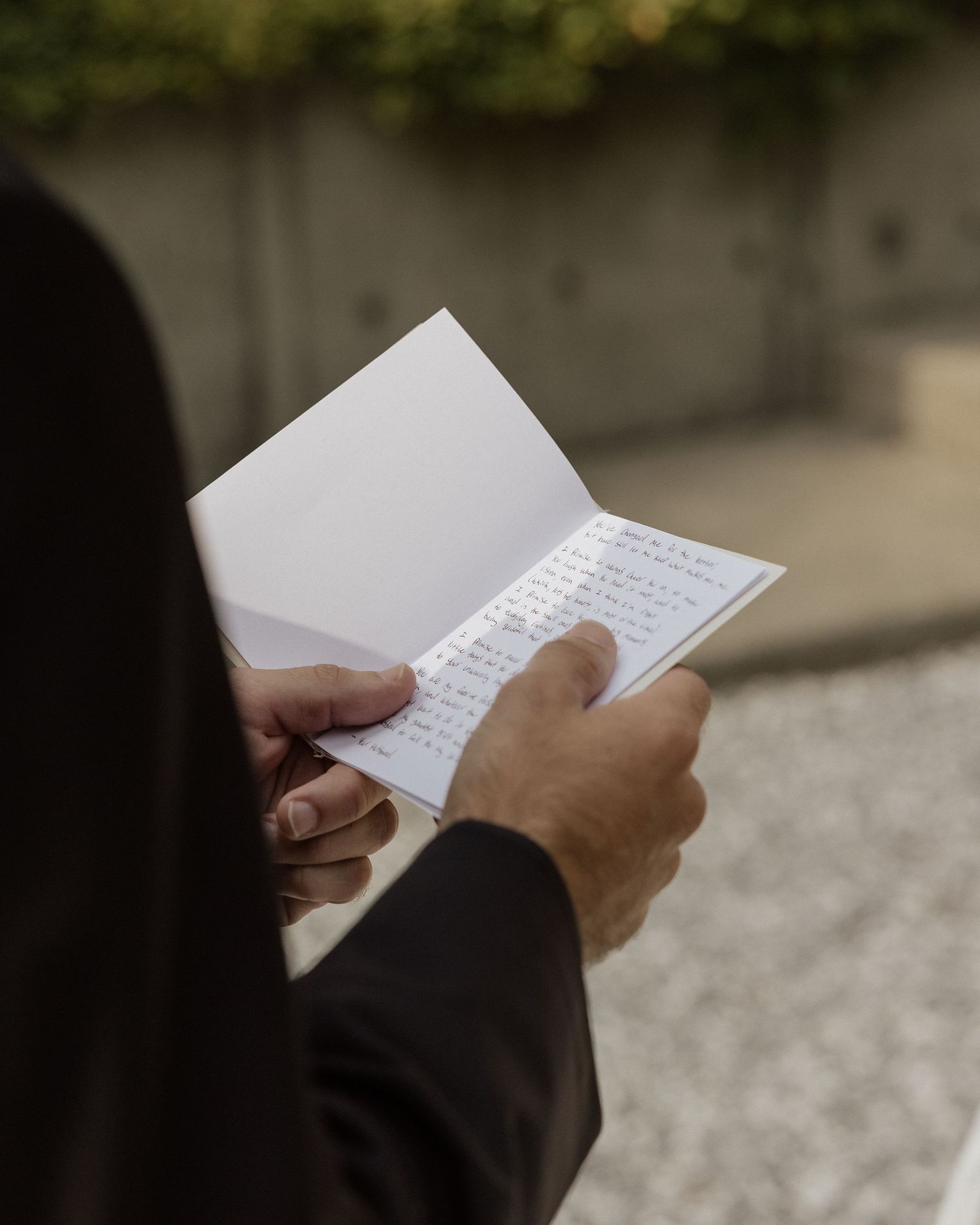 Person in black suit reading handwritten wedding vows from a small white booklet outdoors at a garden ceremony.
