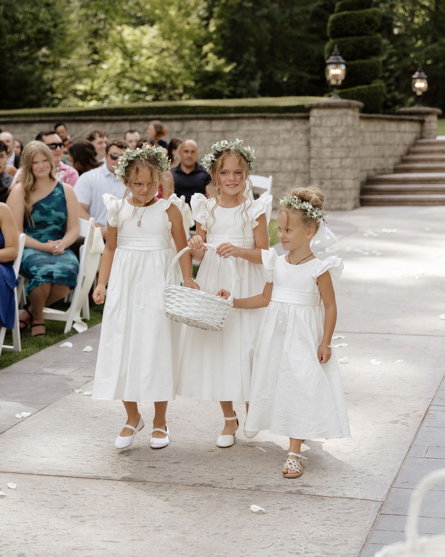 Three flower girls in white dresses and floral crowns walk down a wedding aisle carrying a basket.