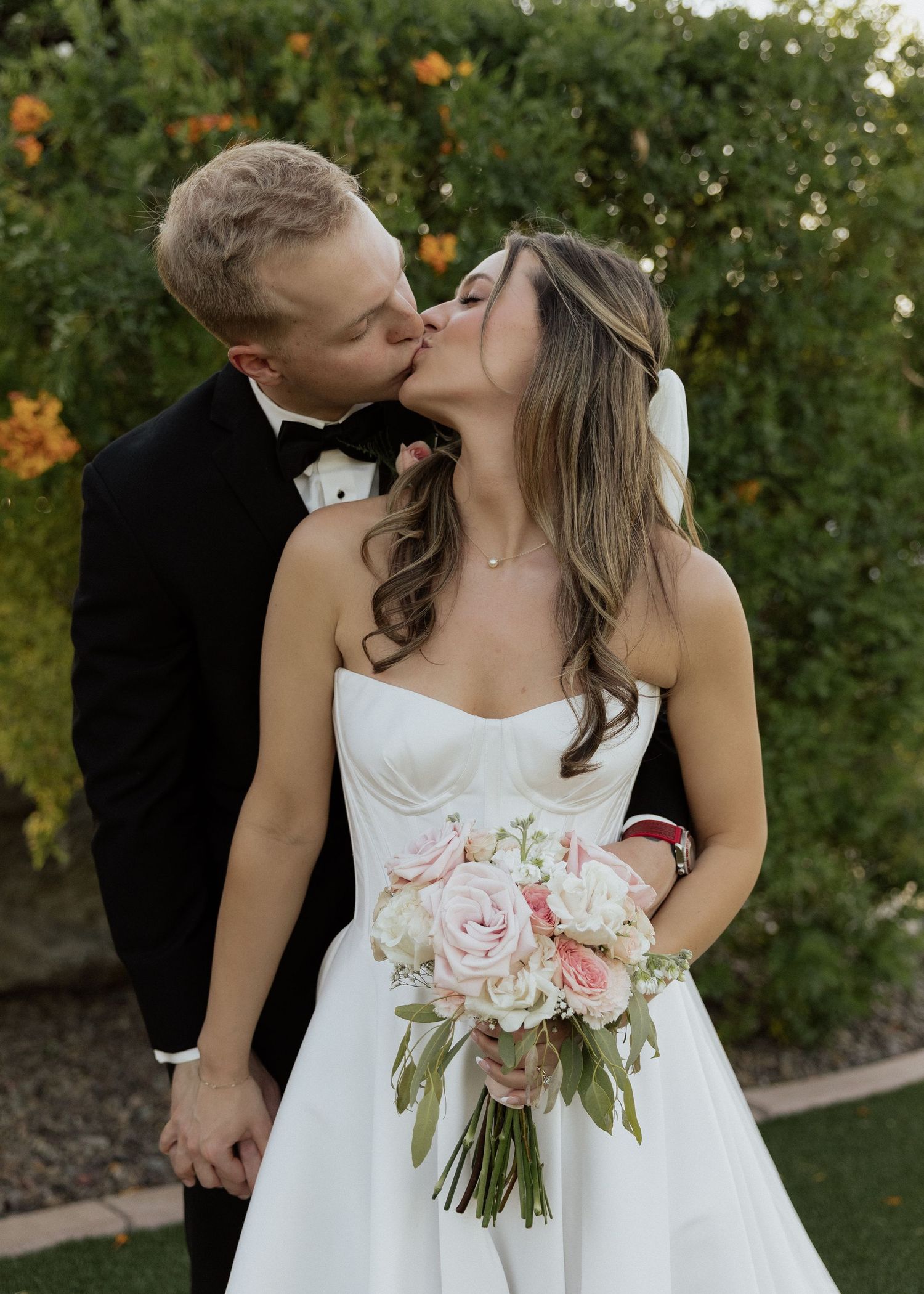 Bride and groom sharing a romantic kiss outdoors, bride holding pink and white floral bouquet in white strapless gown.