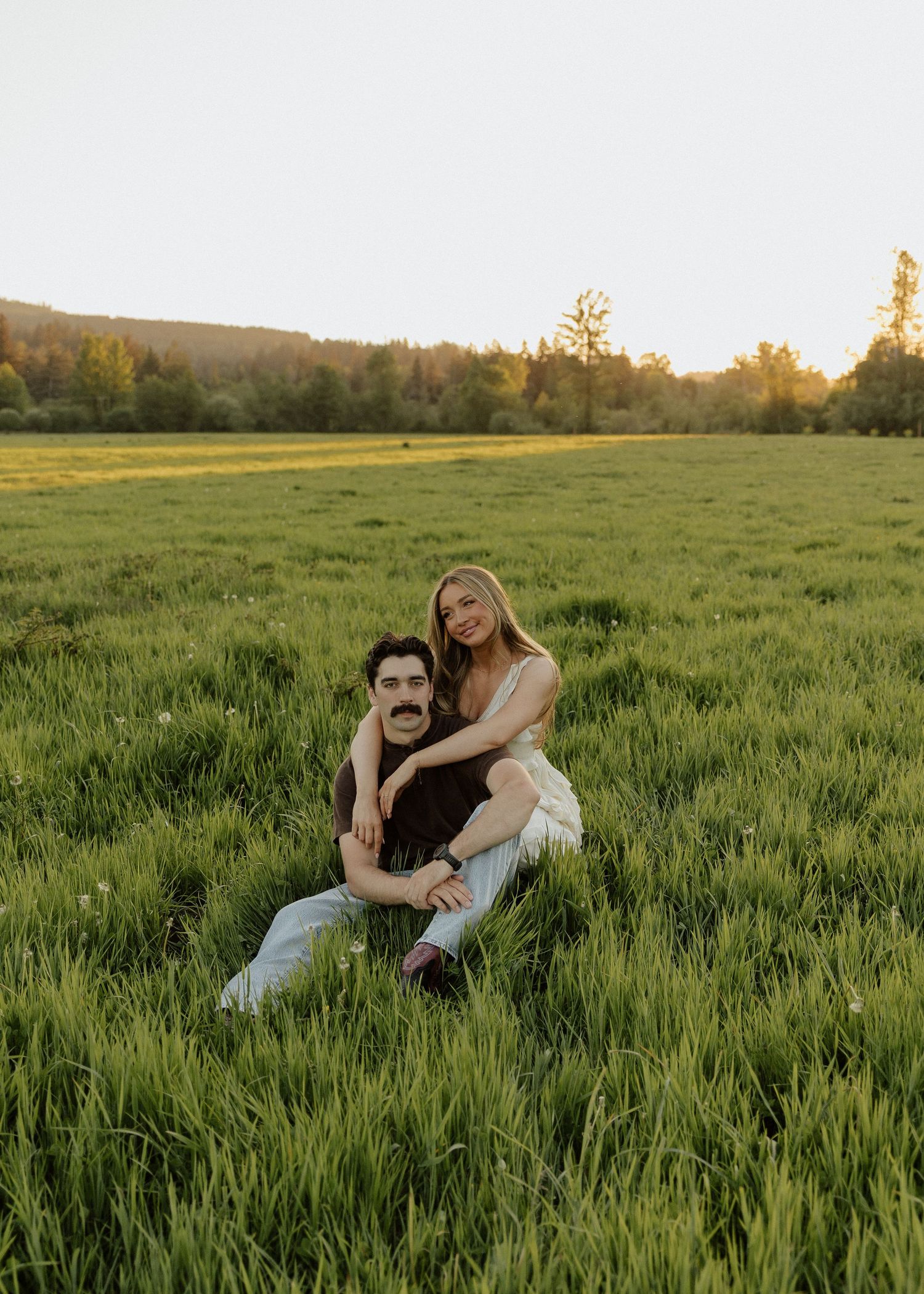A couple sits together in a lush green field at sunset, smiling warmly at the camera.