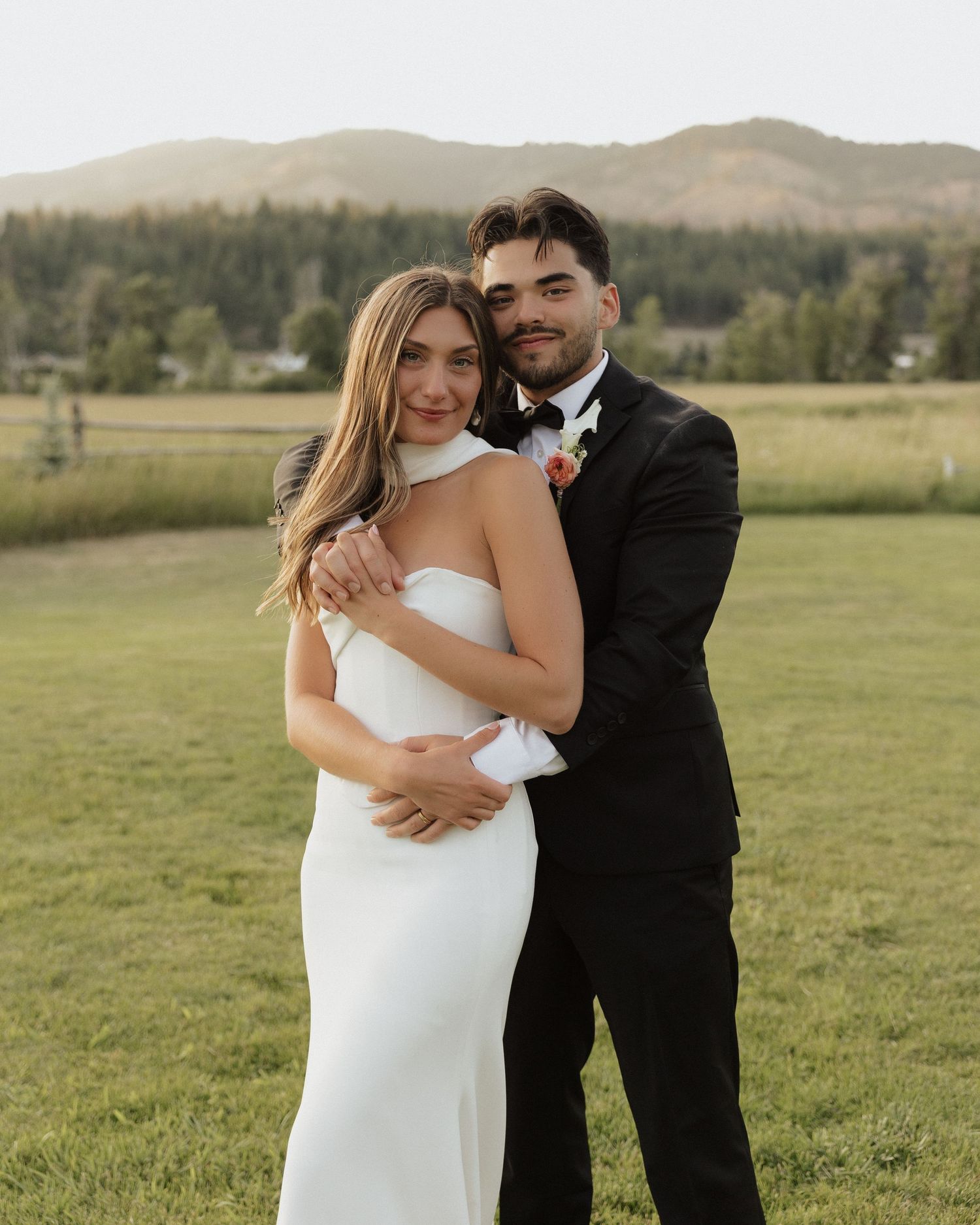 Bride and groom pose together in white wedding dress and black suit with mountain backdrop at sunset.