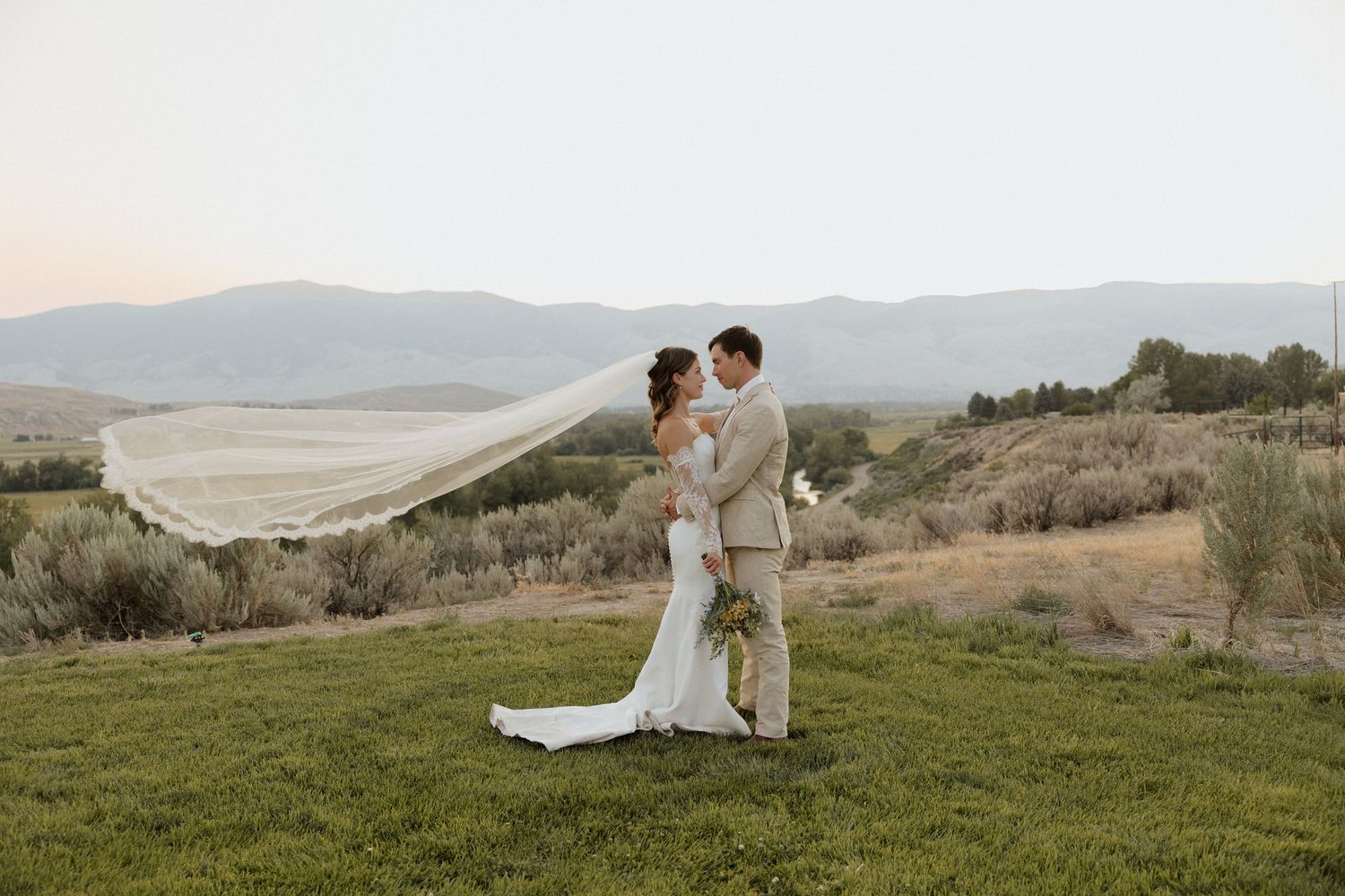Bride and groom embrace on hilltop with flowing veil against mountain landscape.