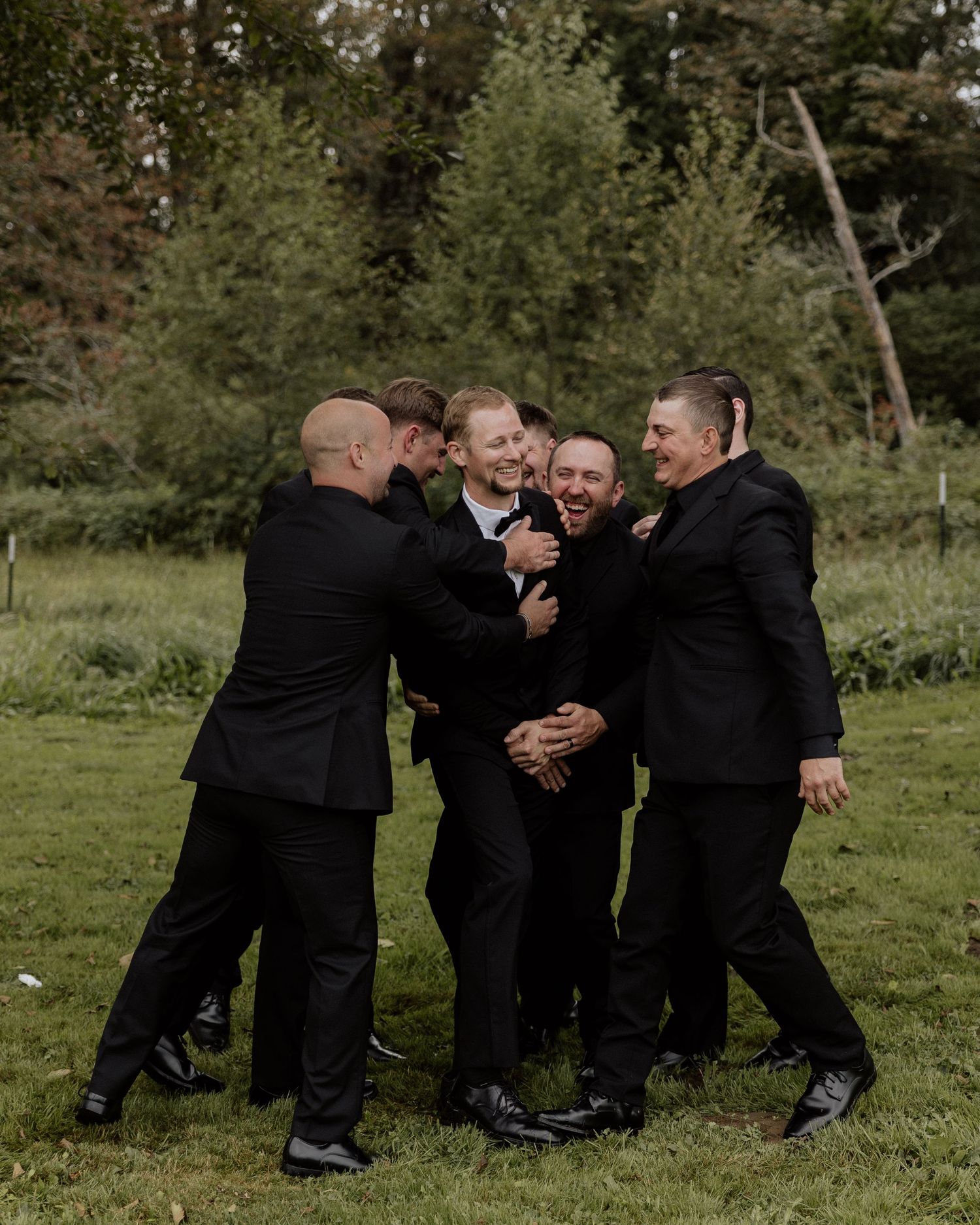 Groomsmen in black suits share a lighthearted moment hugging and laughing together outdoors.