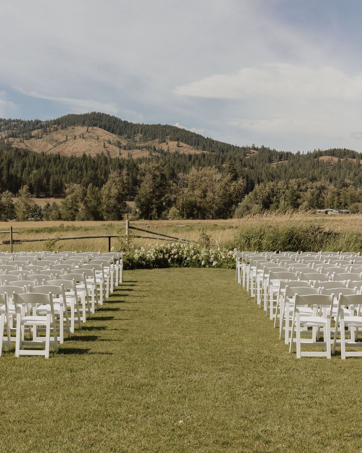 White wooden chairs arranged in rows on a lush green lawn for an outdoor wedding ceremony with mountains in background.