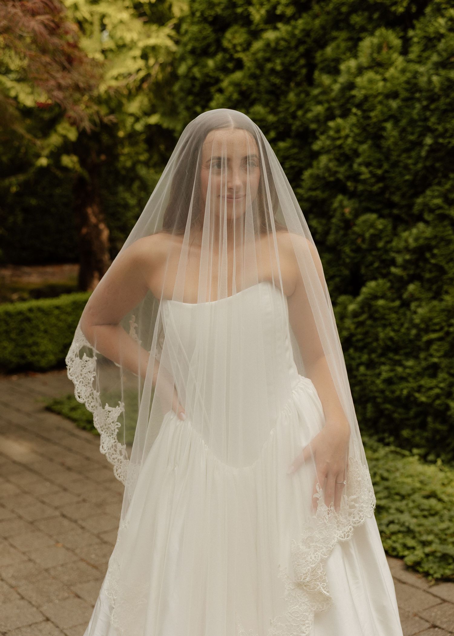 Bride wearing lace-trimmed cathedral veil over strapless white wedding dress outdoors.