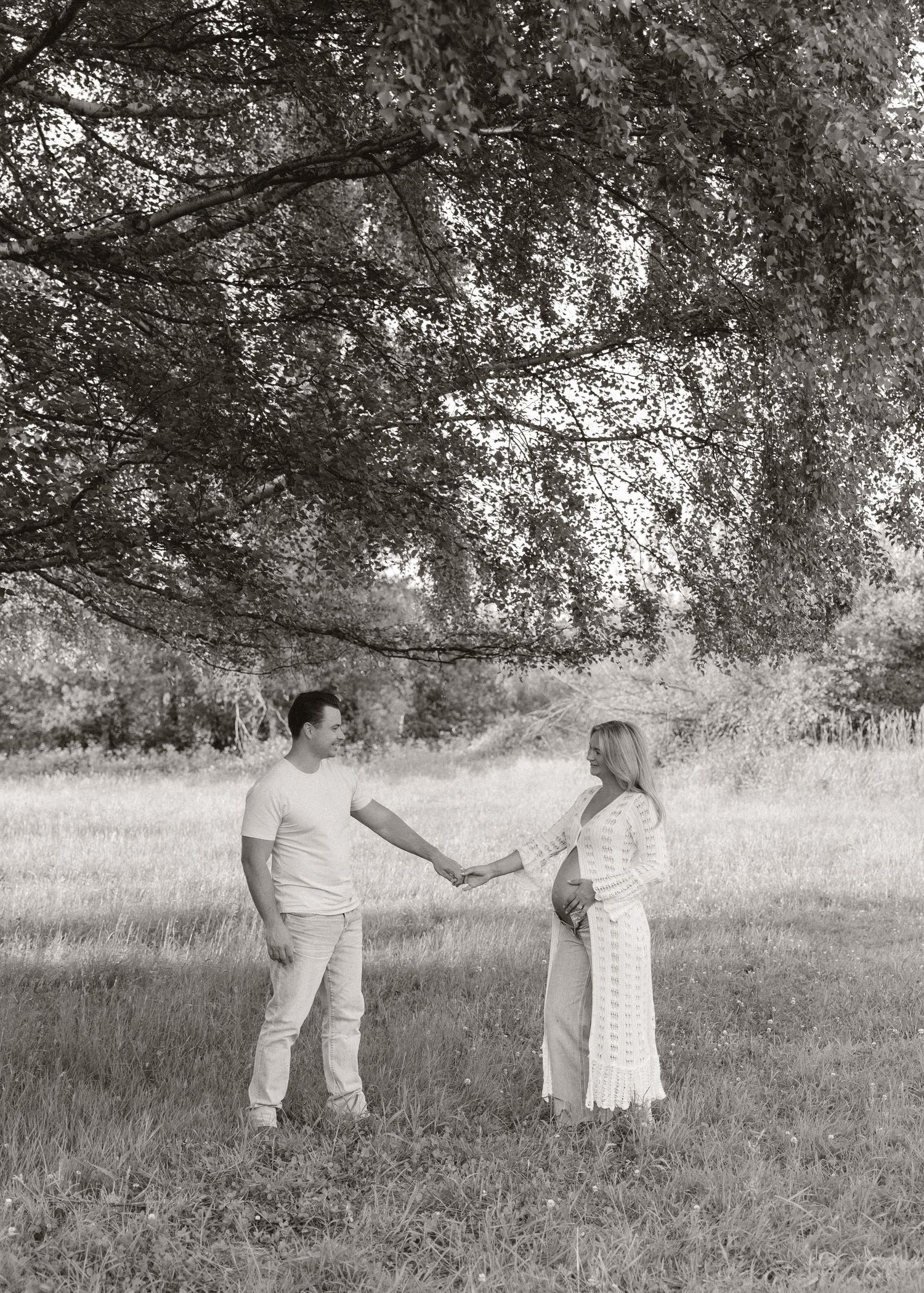 Black and white photo of a couple holding hands in a field under a large tree, woman in white dress appears pregnant.