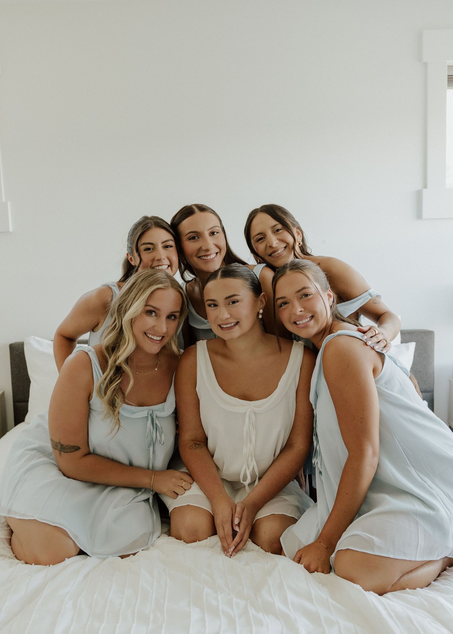 Five smiling women in matching pastel pajamas sitting together on a white bed in a bright, airy room.