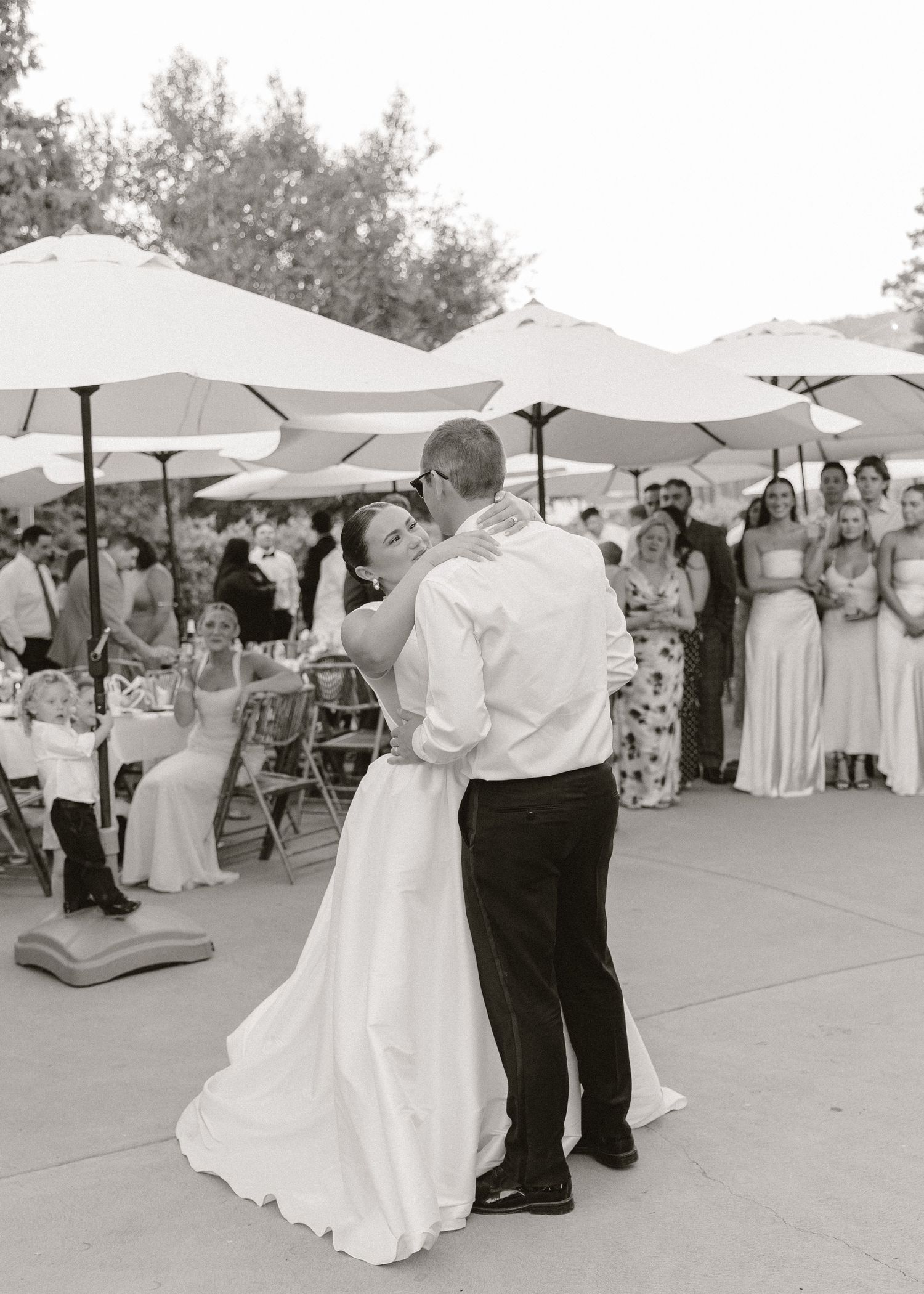 Black and white photo of a bride and groom sharing their first dance outdoors, with guests watching under white umbrellas.