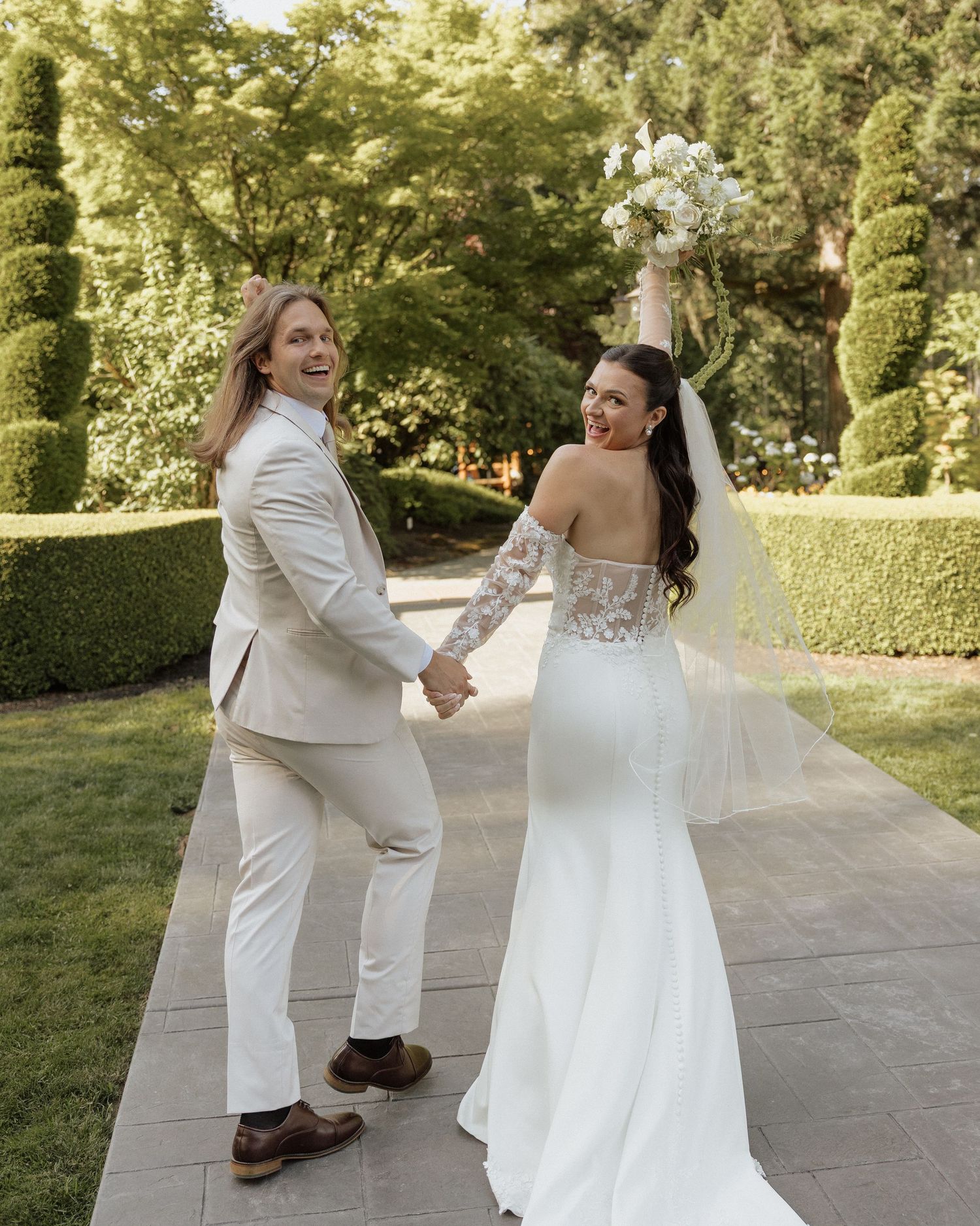Joyful newlyweds walking hand-in-hand, bride raising bouquet triumphantly in garden setting.