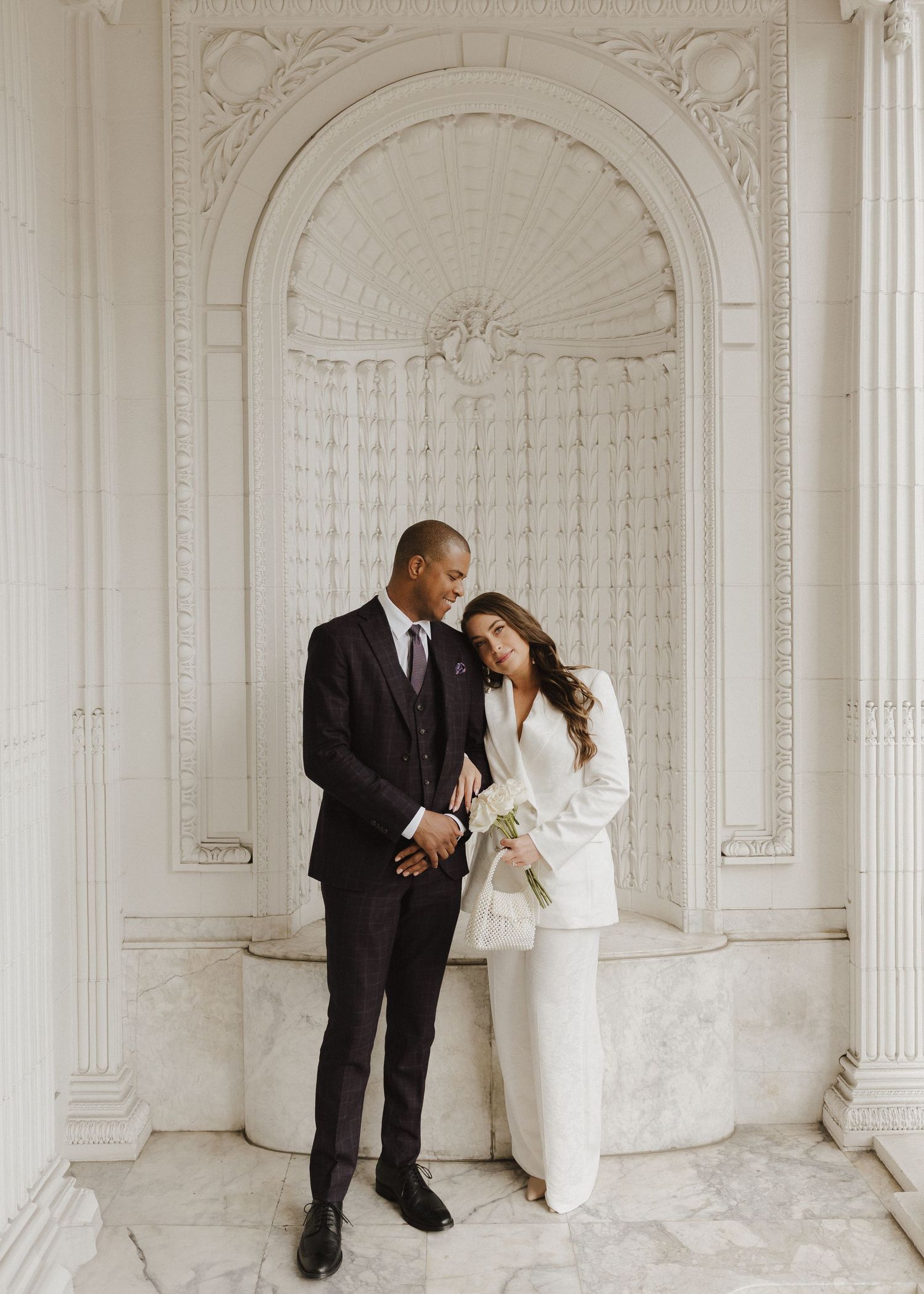 Elegant couple in wedding attire posing in a white marble archway, groom in dark suit, bride in white pantsuit holding bouquet.