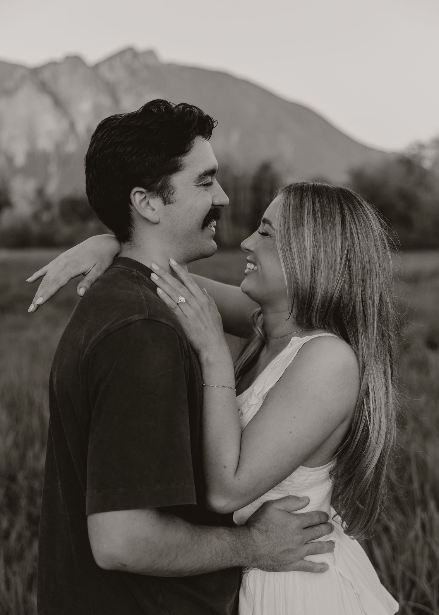 Black and white photo of a smiling couple embracing in a field with a mountain in the background.