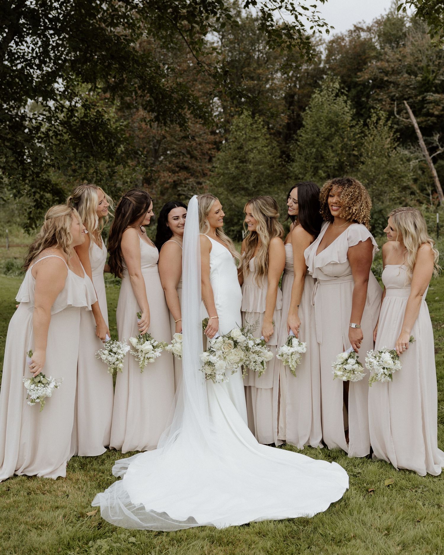 Wedding party stands in a garden setting with bridesmaids in light neutral dresses holding white bouquets.