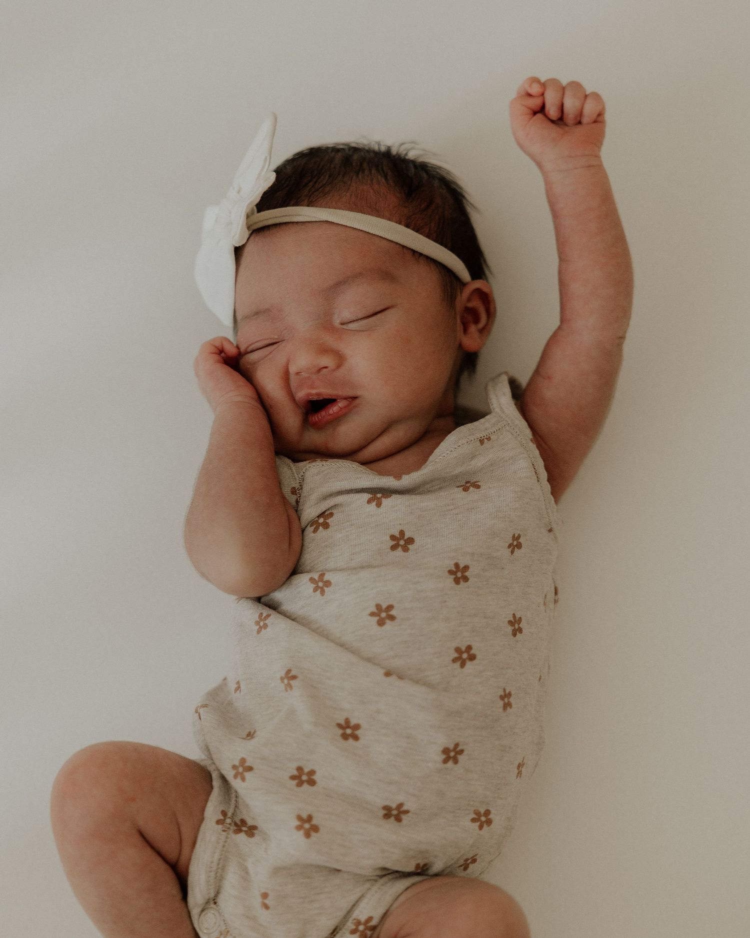 Peaceful sleeping newborn wearing a floral onesie and white headband stretches an arm upward.