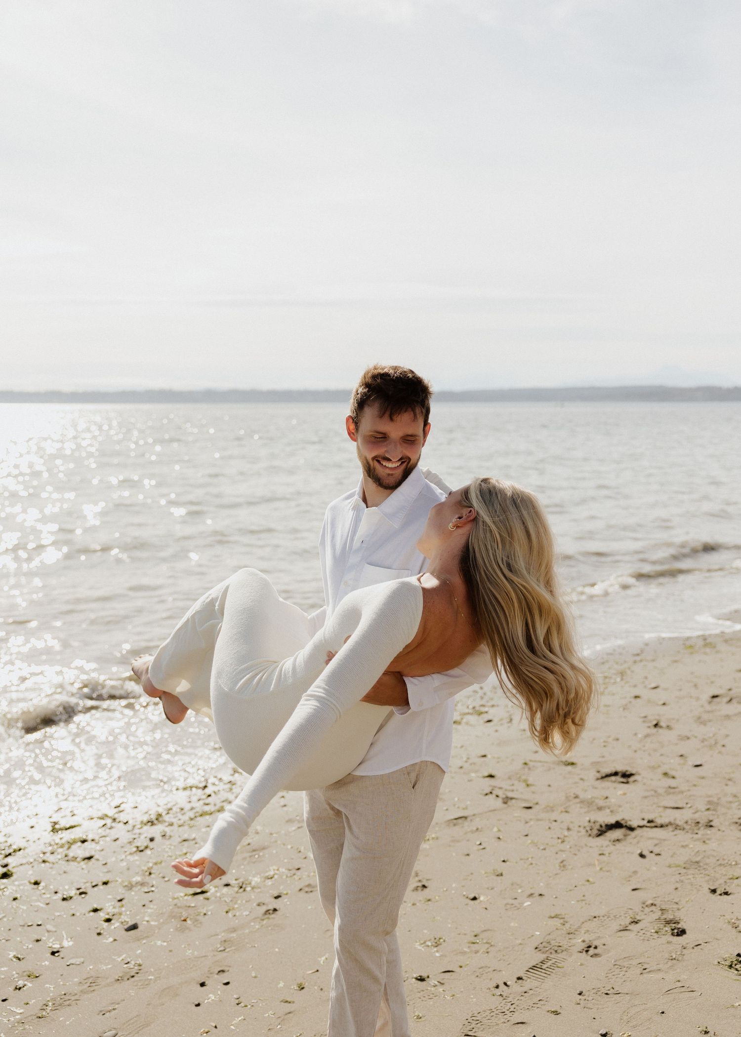 A smiling man in white carries a laughing blonde woman on a sunny beach near calm water.