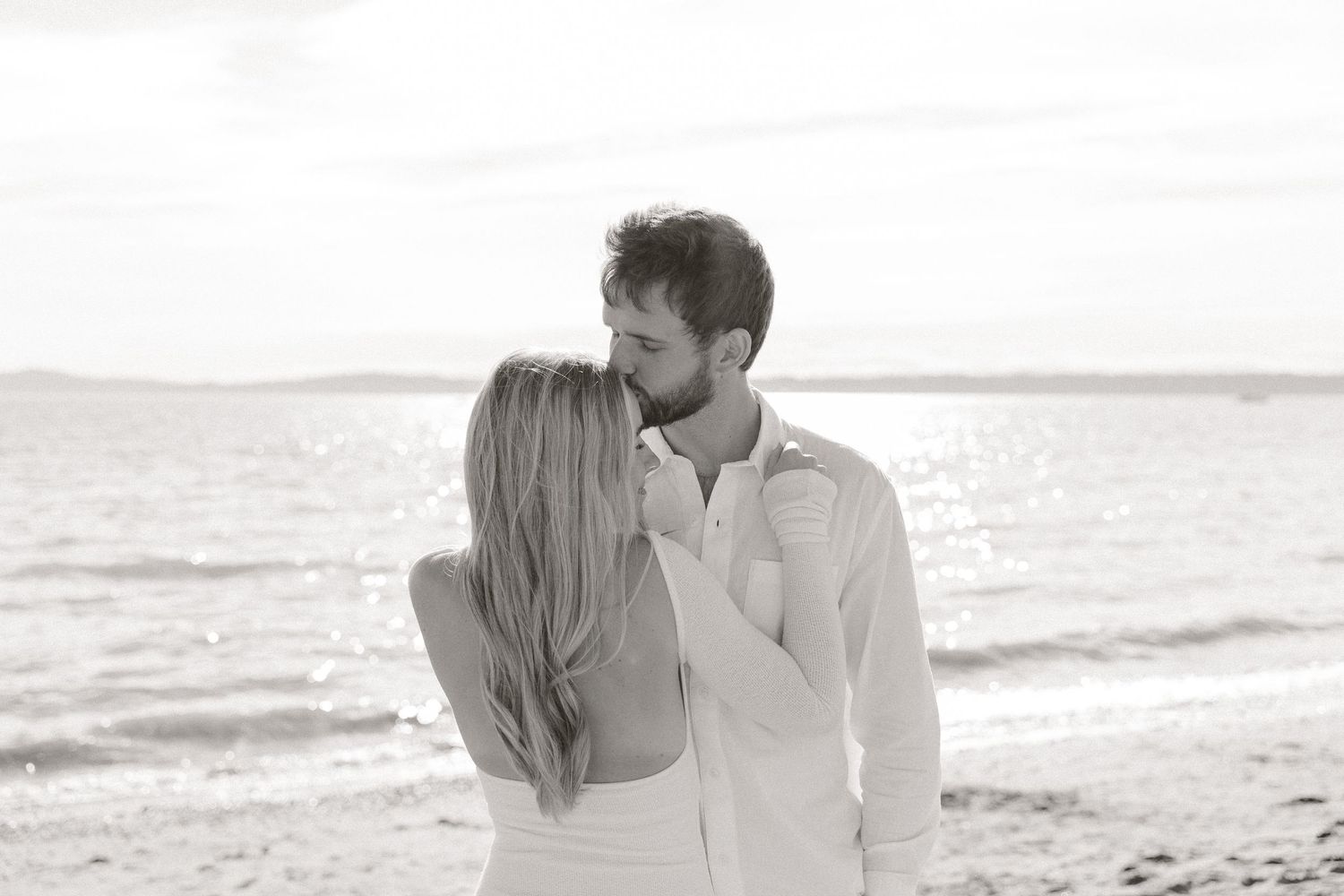Romantic couple sharing a tender moment on a sunlit beach with sparkling ocean behind them.