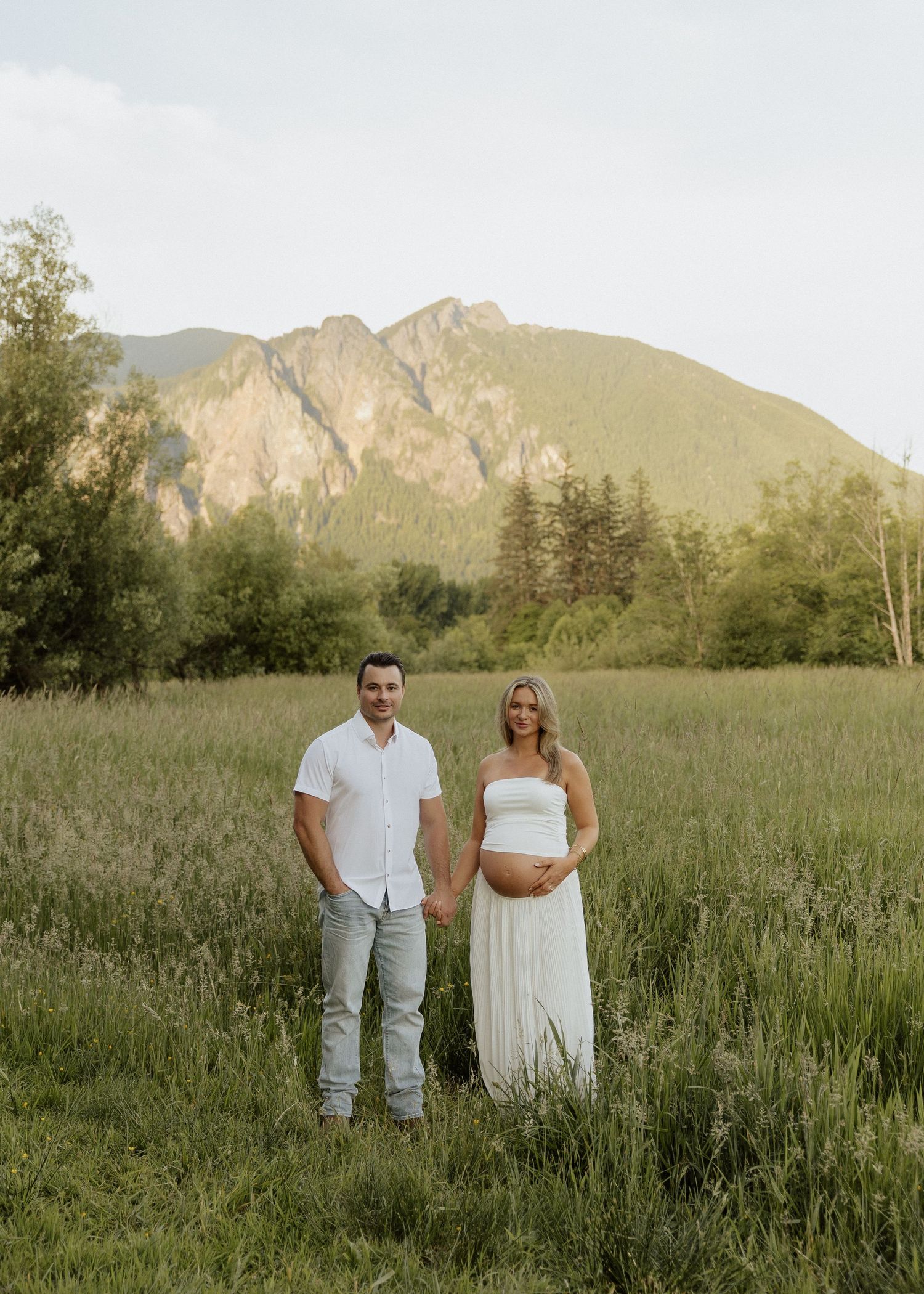 Couple posing in a lush green meadow with mountain backdrop during golden hour maternity photoshoot.