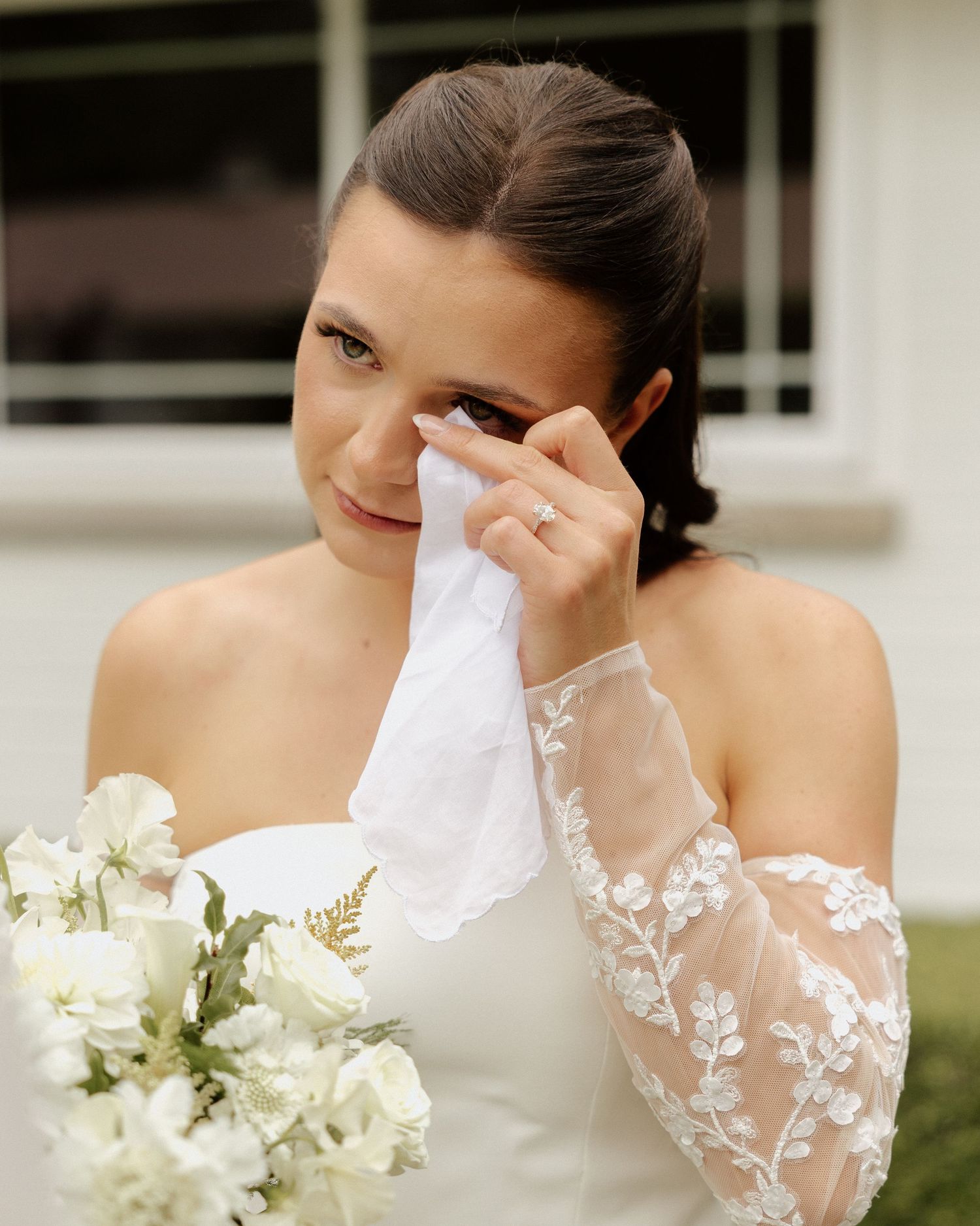 Tearful bride in white off-shoulder gown dabbing eyes with handkerchief holding floral bouquet.