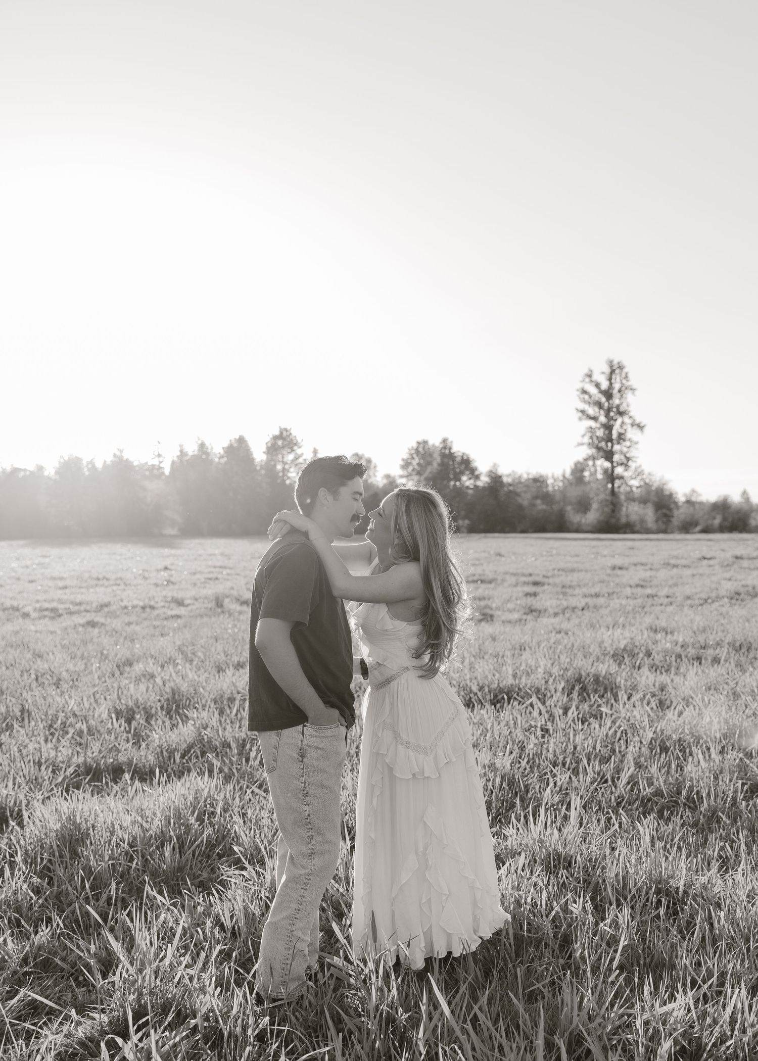 Black and white photo of a romantic couple sharing a tender moment in a sunlit open field with trees in the background.