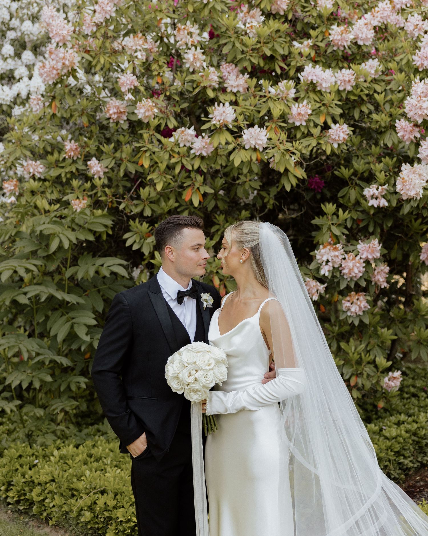 Bride and groom share a romantic moment in front of blooming pink flowers, bride wearing white gown, veil, and gloves.