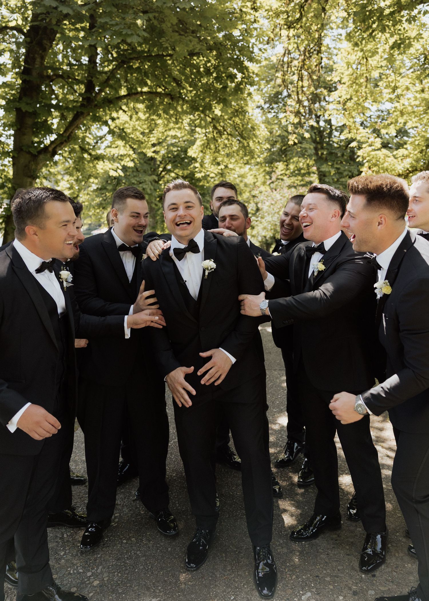 Groom and groomsmen laughing together outdoors in black tuxedos with bow ties and boutonnieres at a wedding celebration.