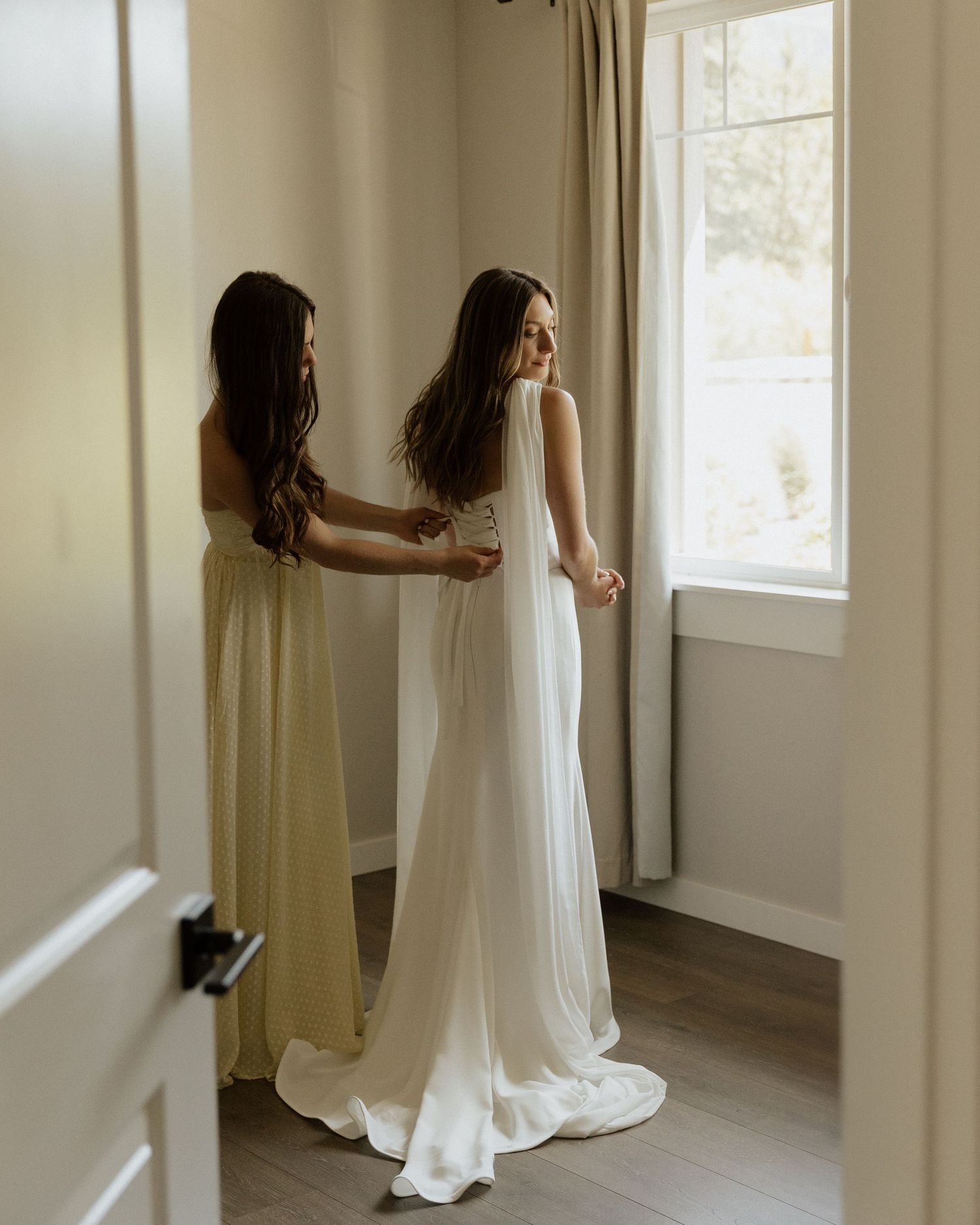 Bridesmaid in yellow dress helping bride in white gown with veil near a bright window in an elegant room.