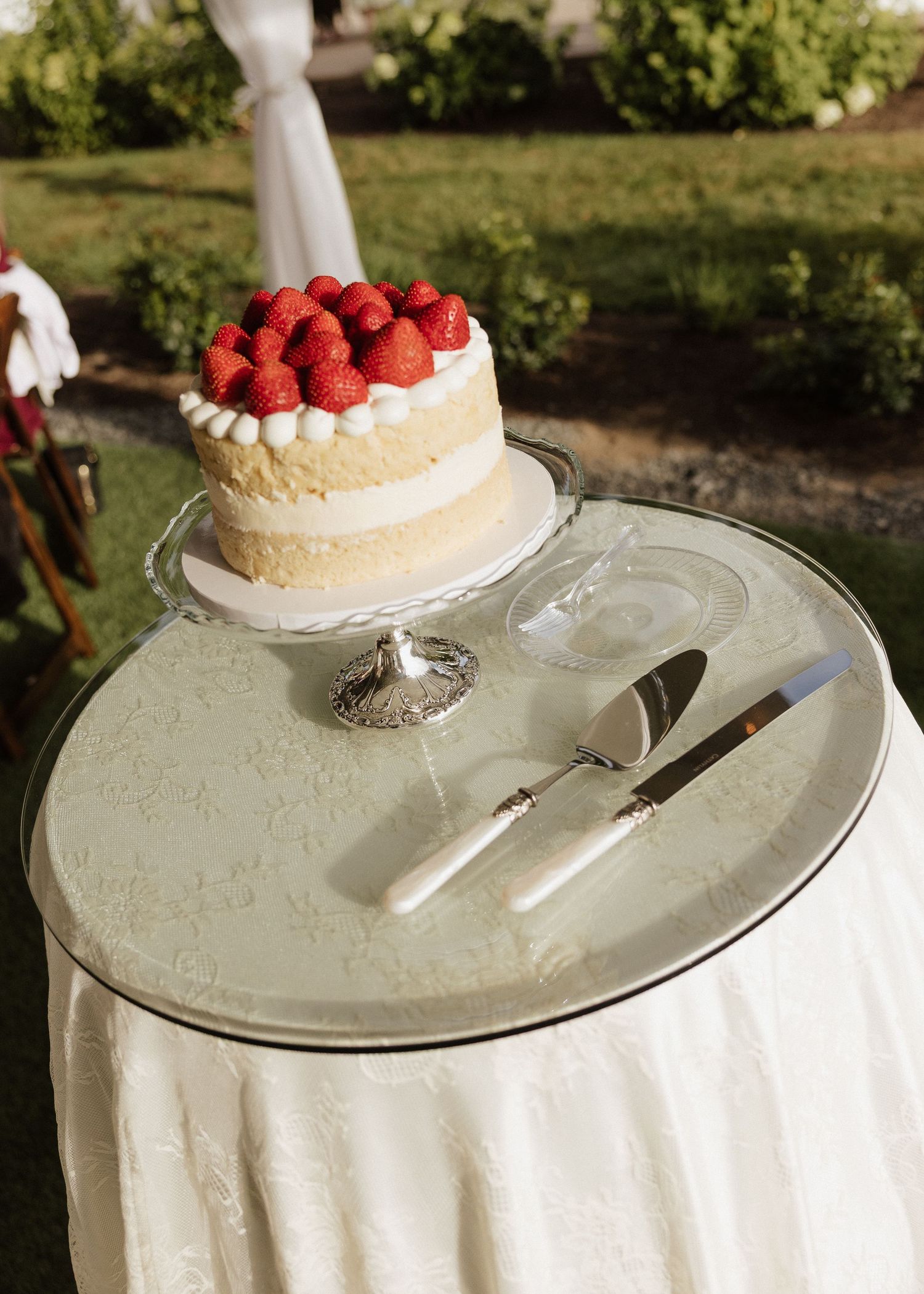 Elegant naked wedding cake topped with fresh strawberries displayed on a silver cake stand outdoors.