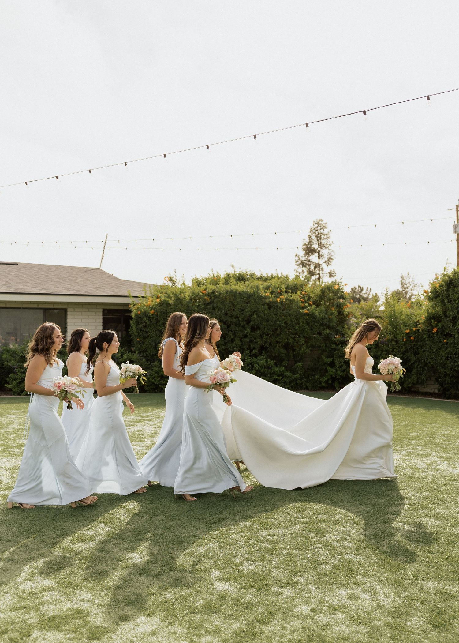 Bridesmaids in light blue gowns walk across green lawn carrying bride's white wedding dress train outdoors.