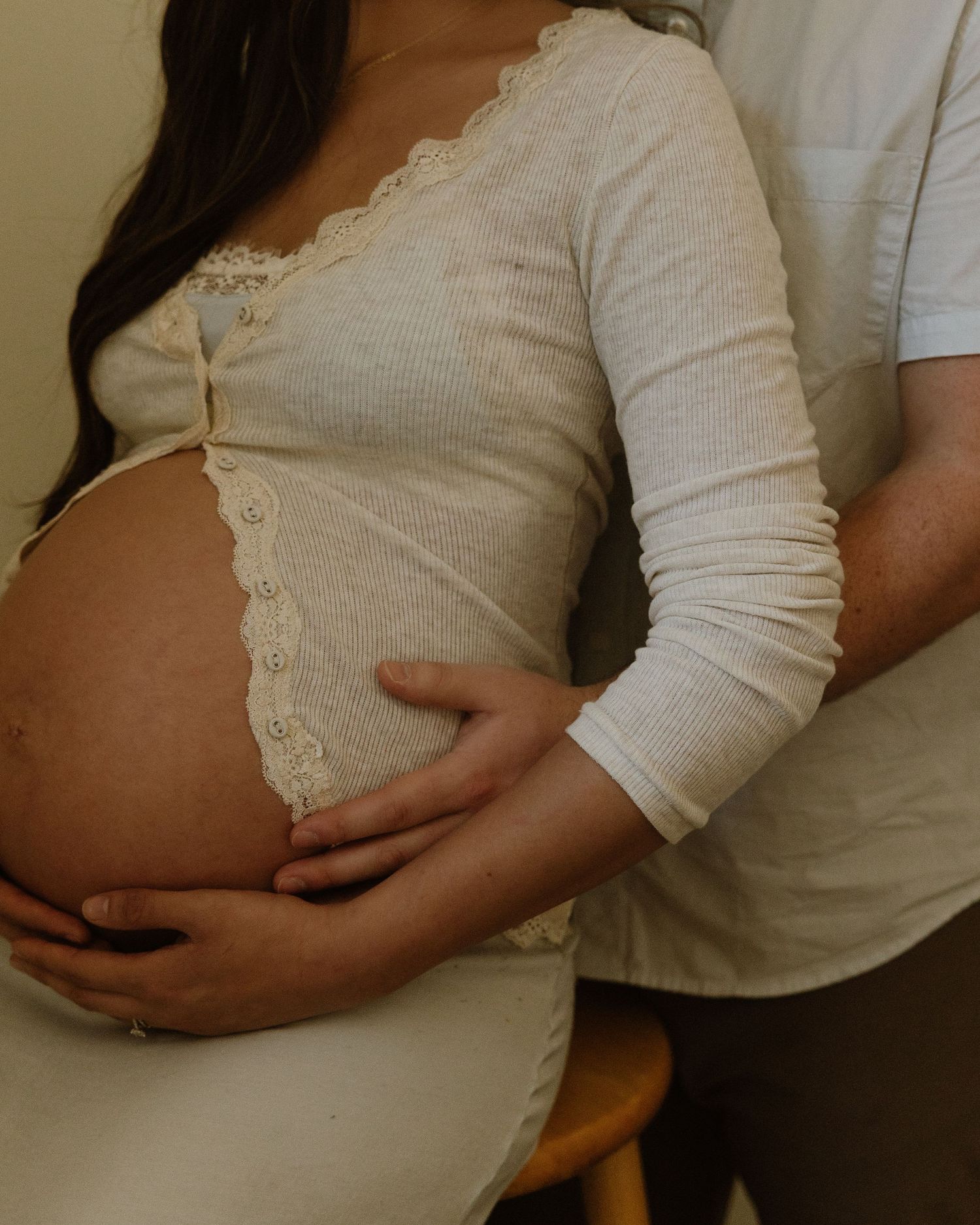 Close-up of hands gently cradling a pregnant belly in a white lace dress.