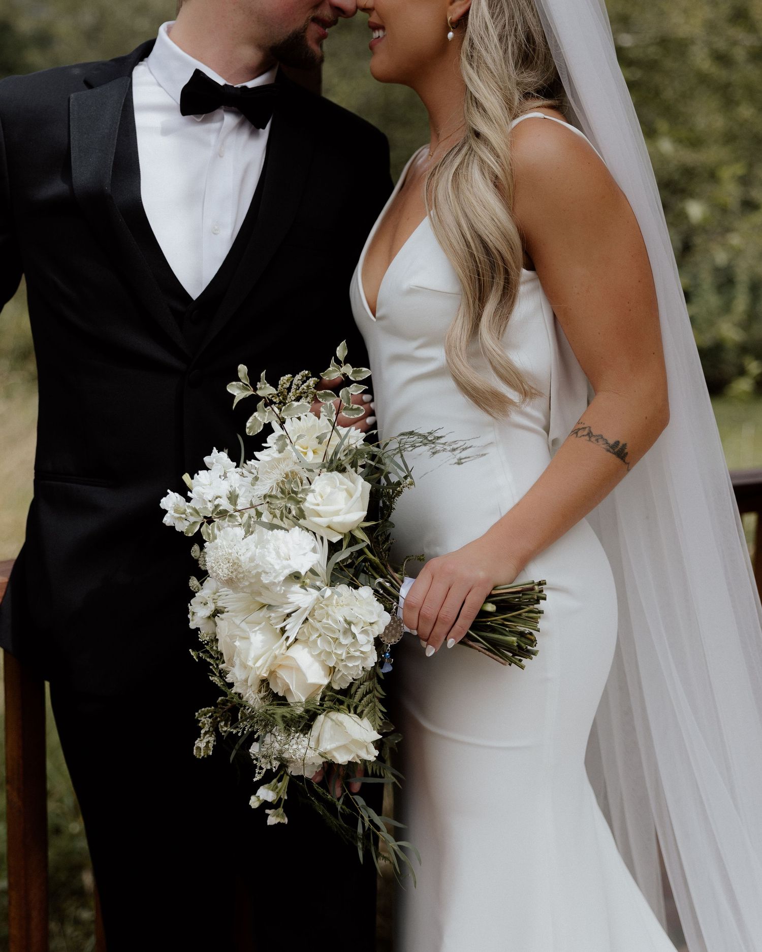 Elegant bride in fitted white dress holds white floral bouquet while embracing groom in black tuxedo.