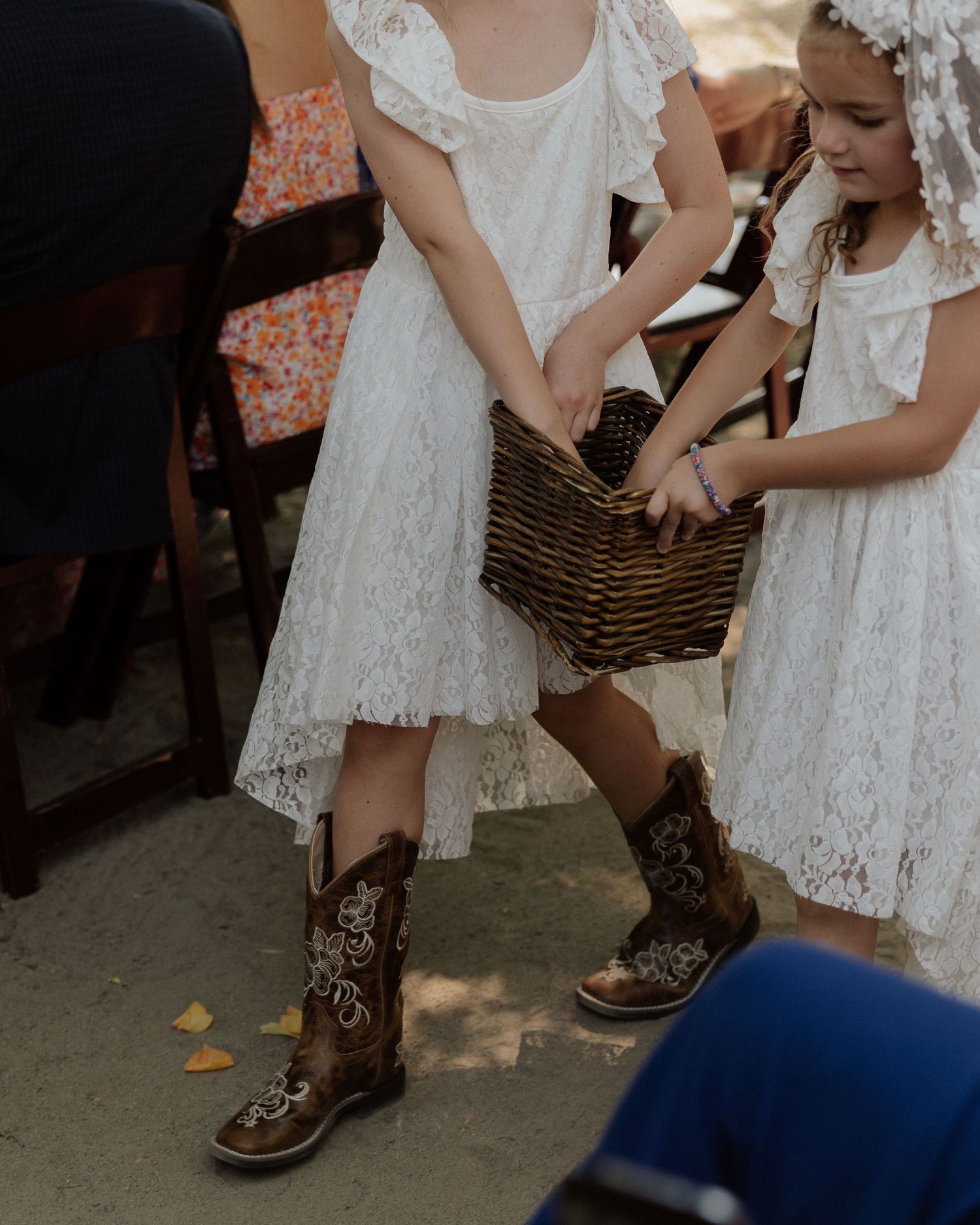 Young flower girls in white lace dresses and cowboy boots carry woven basket at outdoor wedding.