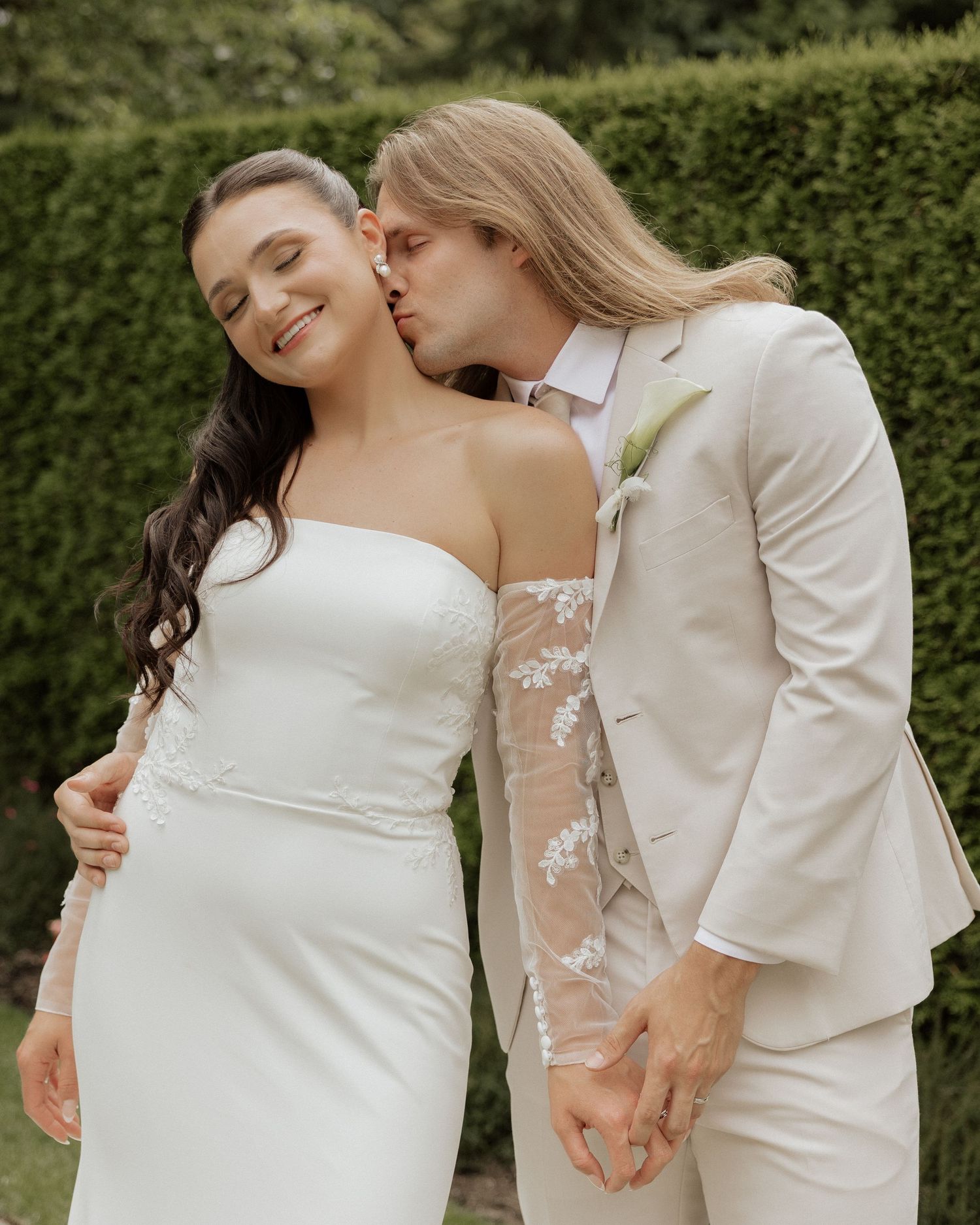 Two people wearing white outfits embrace intimately in a garden setting while sharing tender moments.